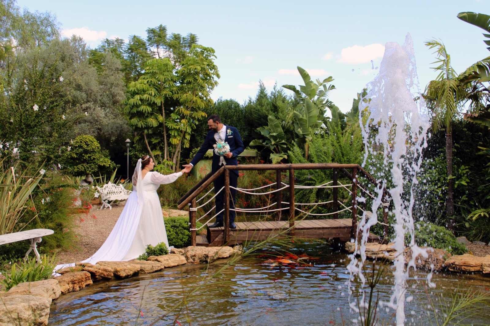 Una novia y un novio, vestidos de novia, posan junto a un estanque con fuente. 