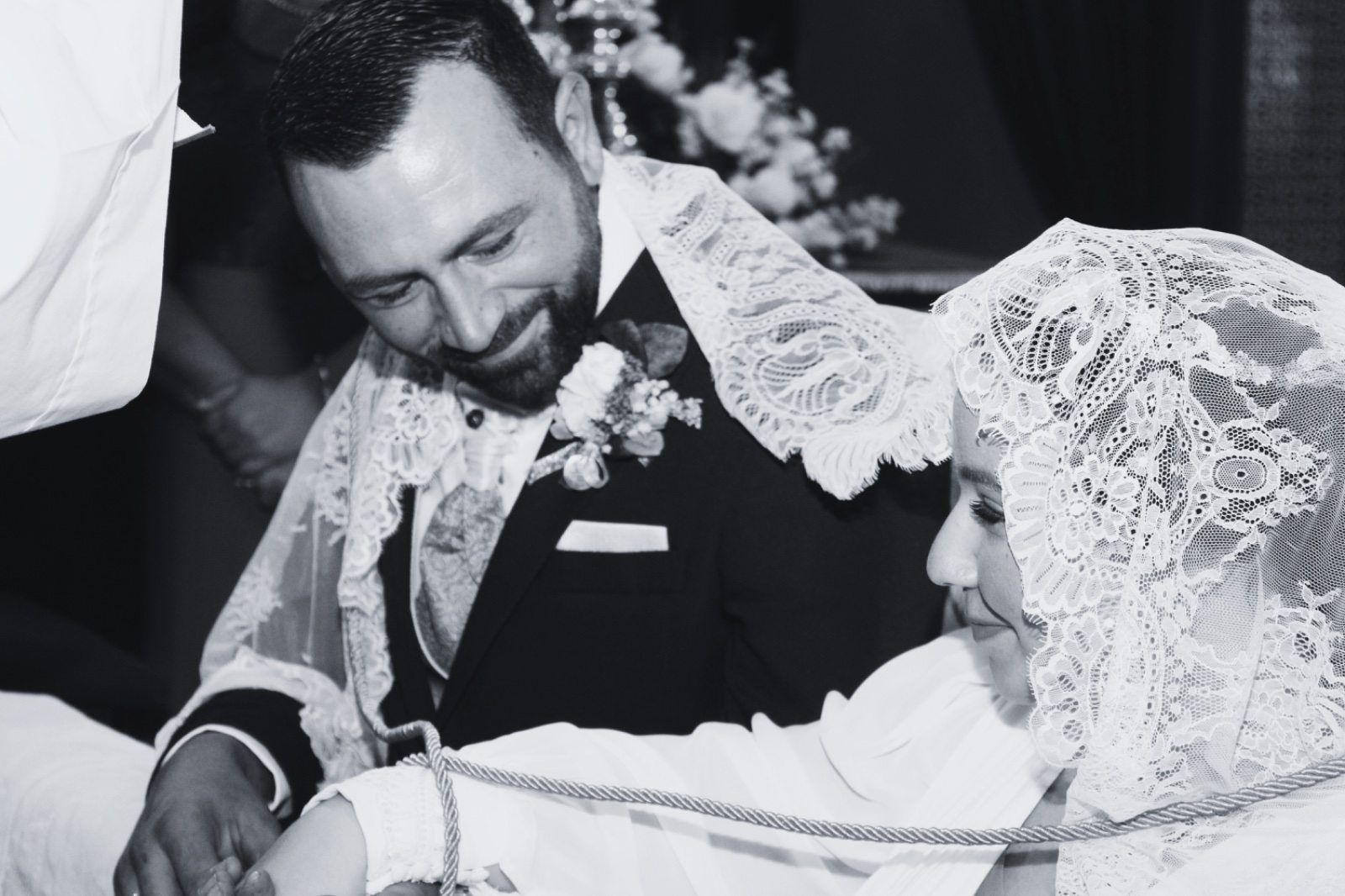 El novio sonríe a la novia mientras se unen con un cordón durante la ceremonia nupcial. La novia lleva un velo de encaje.