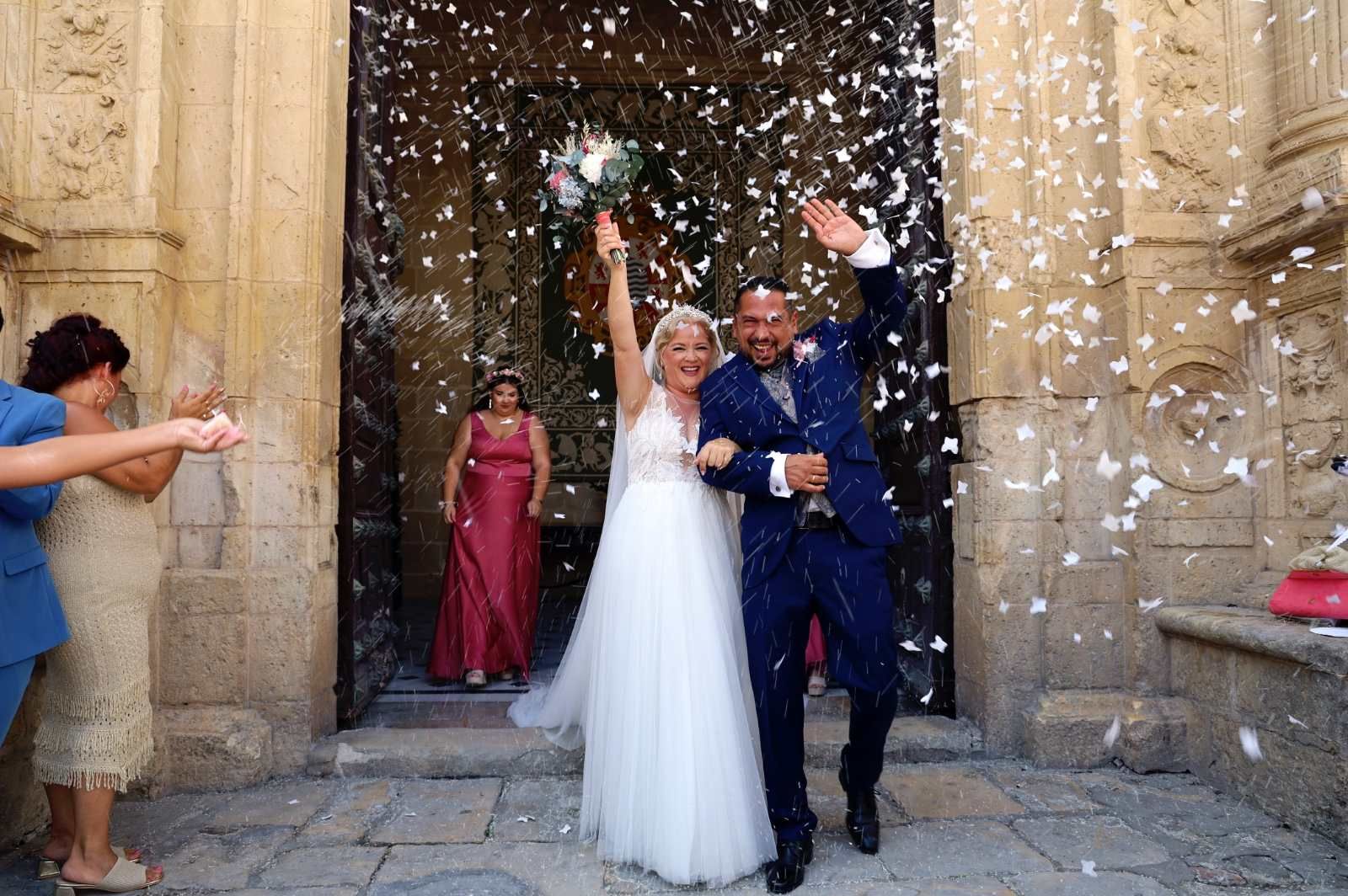 Recién casados saliendo de un edificio de piedra cubierto de confeti.