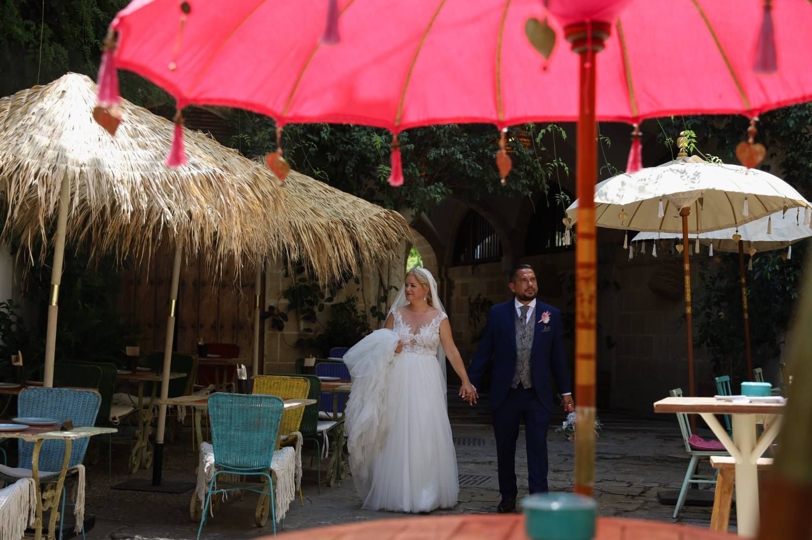 Una pareja de novios se toma de la mano bajo una sombrilla rosa en un colorido escenario al aire libre. 