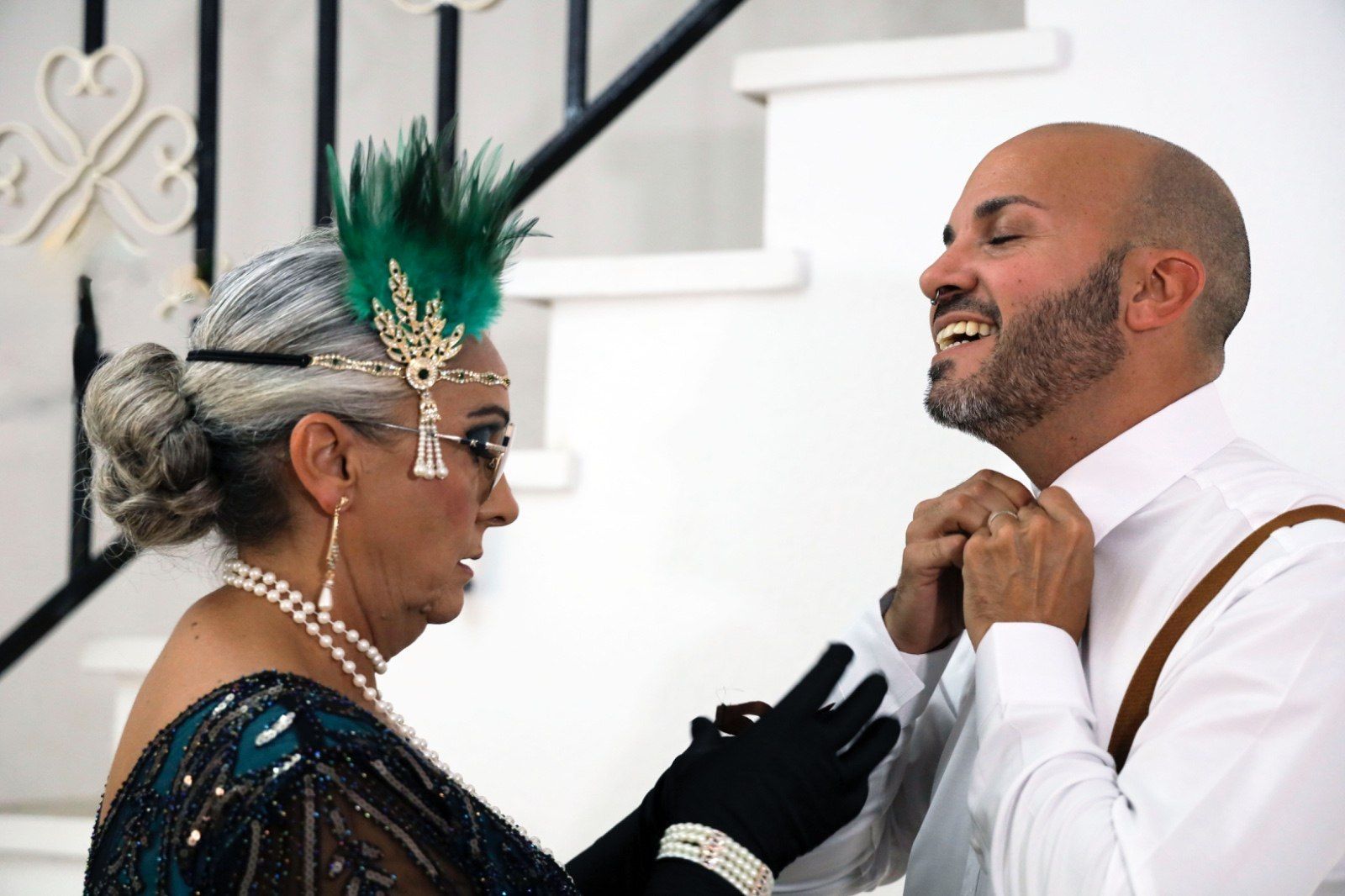 Una mujer con traje de flapper le ajusta la corbata a un hombre. Sonríen en una casa con escalera.