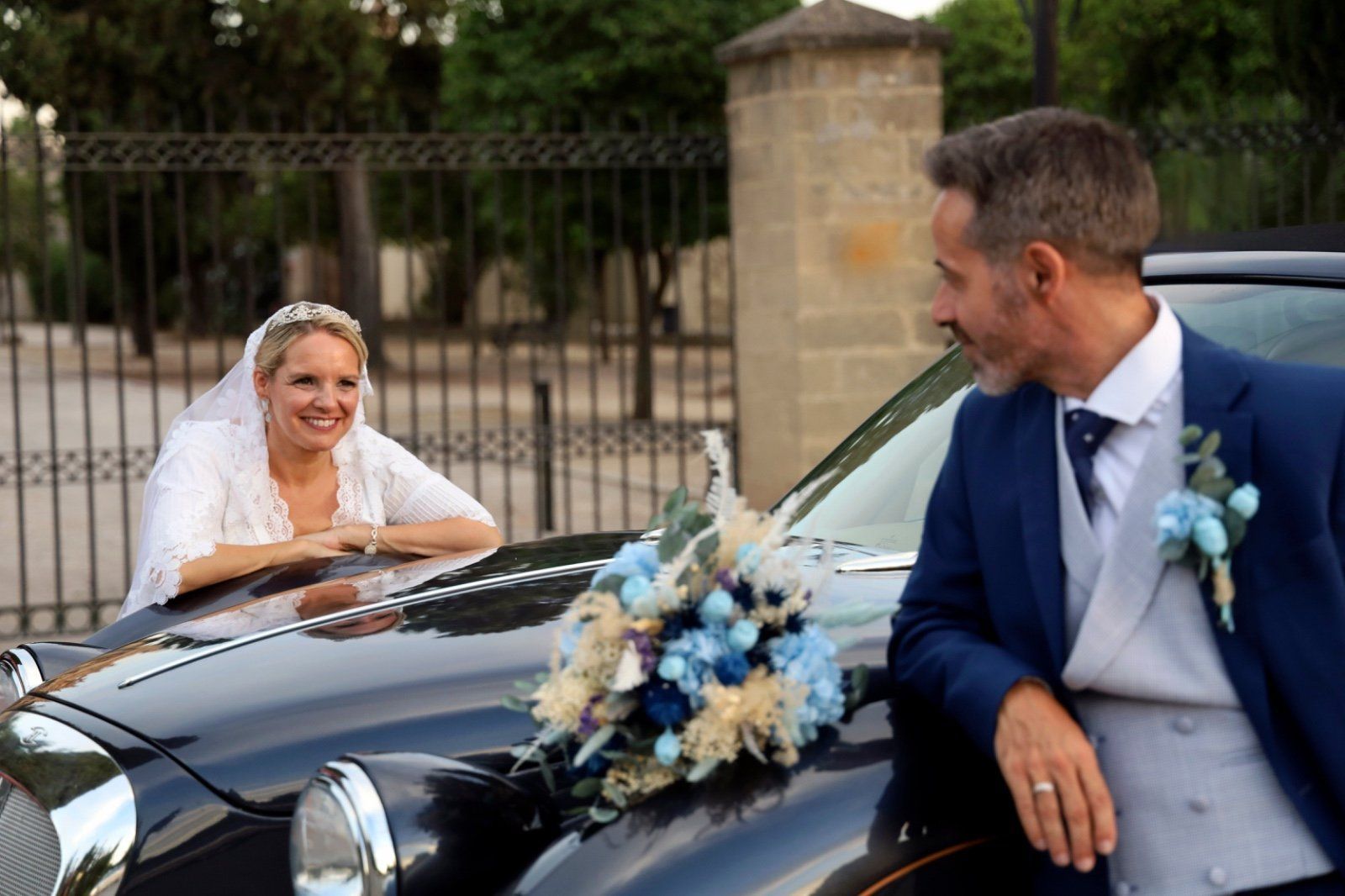Los novios se sonríen, apoyados en un coche negro clásico.