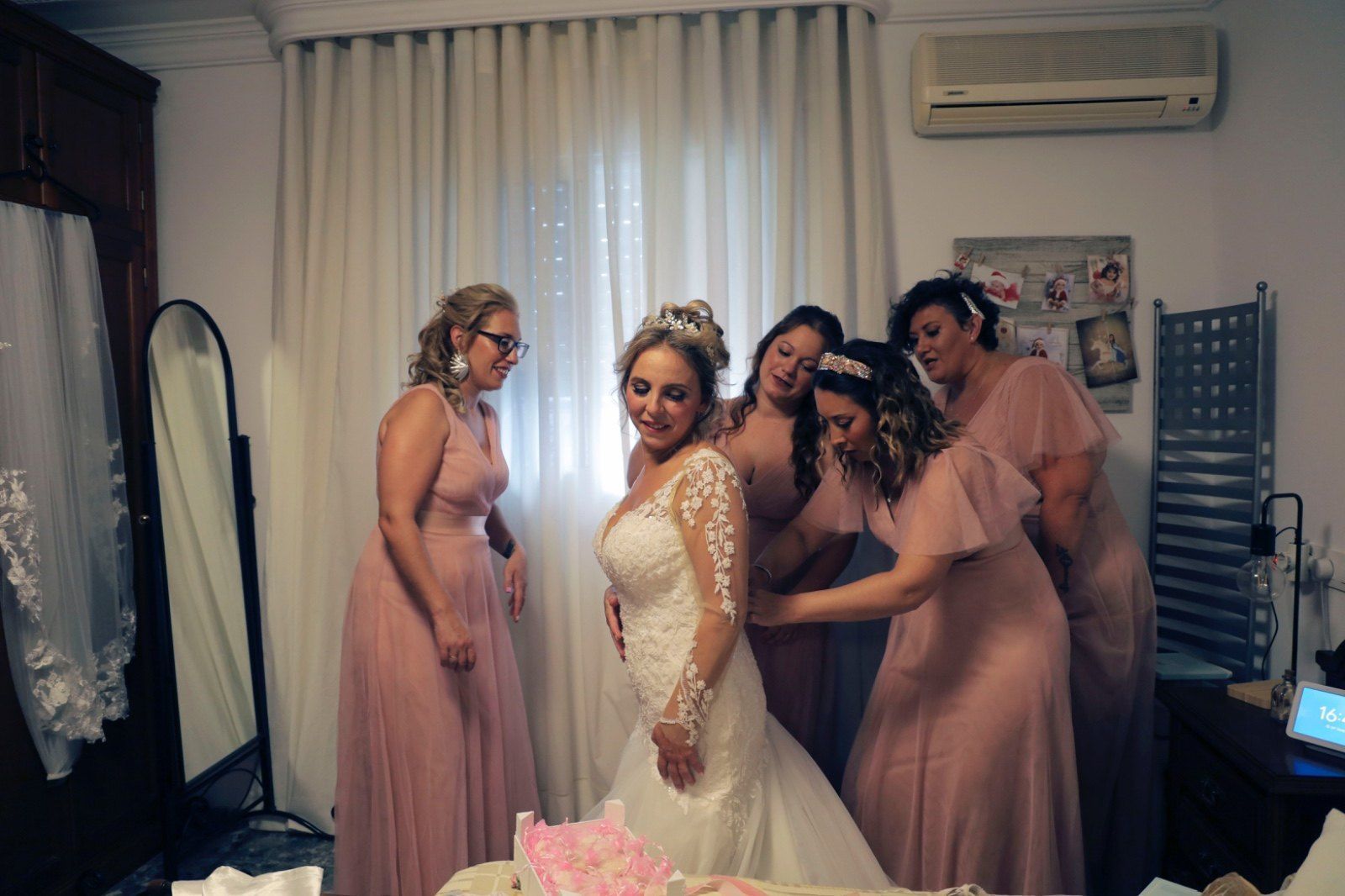 La novia con un vestido blanco con mangas de encaje es ayudada por damas de honor con vestidos rosas.