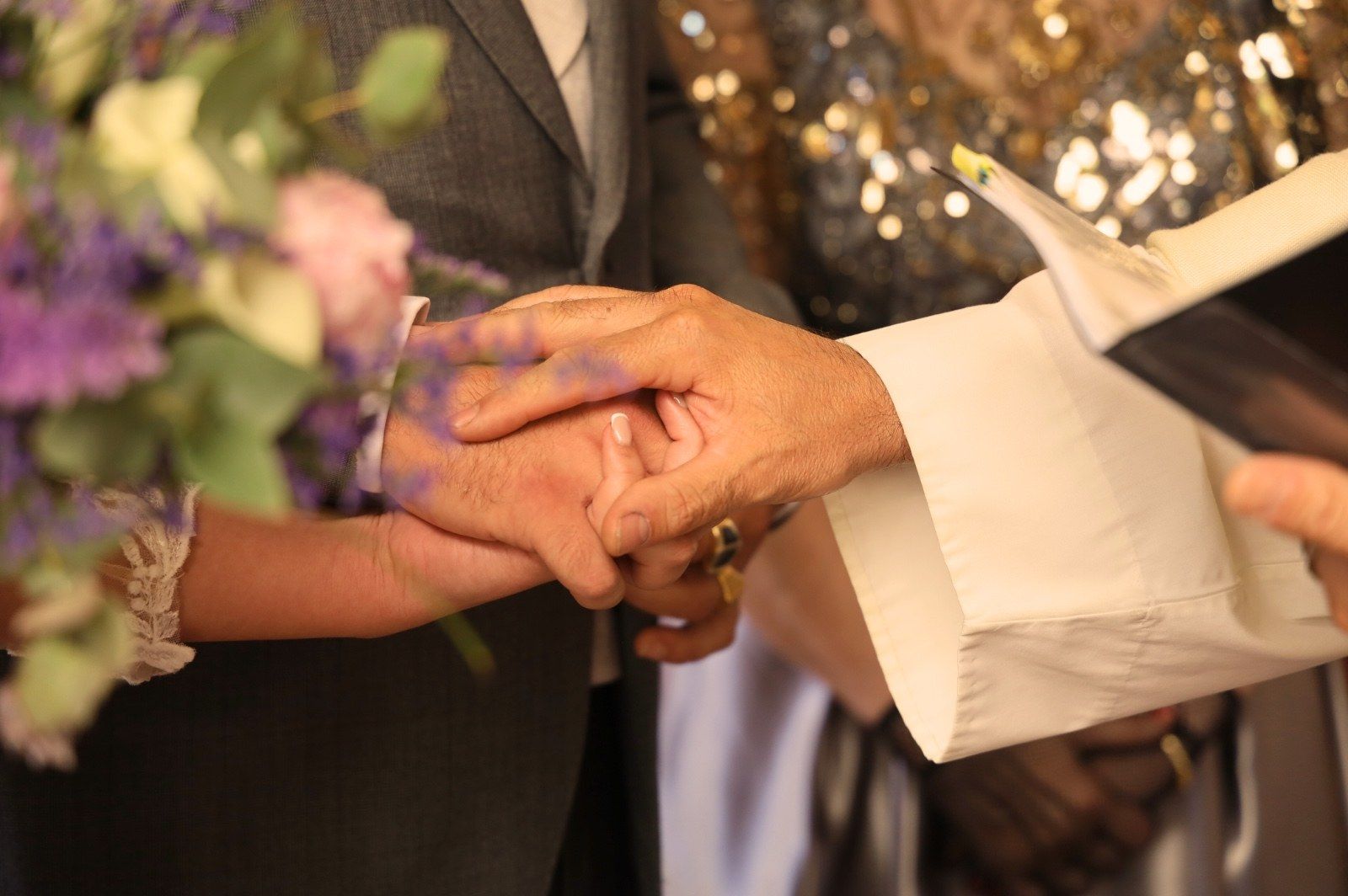 Manos de una pareja abrazándose durante una ceremonia de boda, con la mano de un ministro encima y un ramo de flores visible.