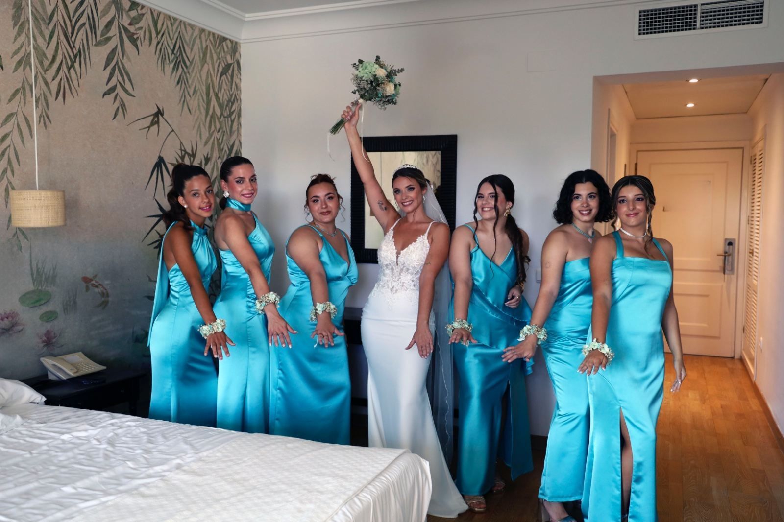 Una novia con vestido blanco y damas de honor con vestidos turquesa posan en una habitación de hotel. 