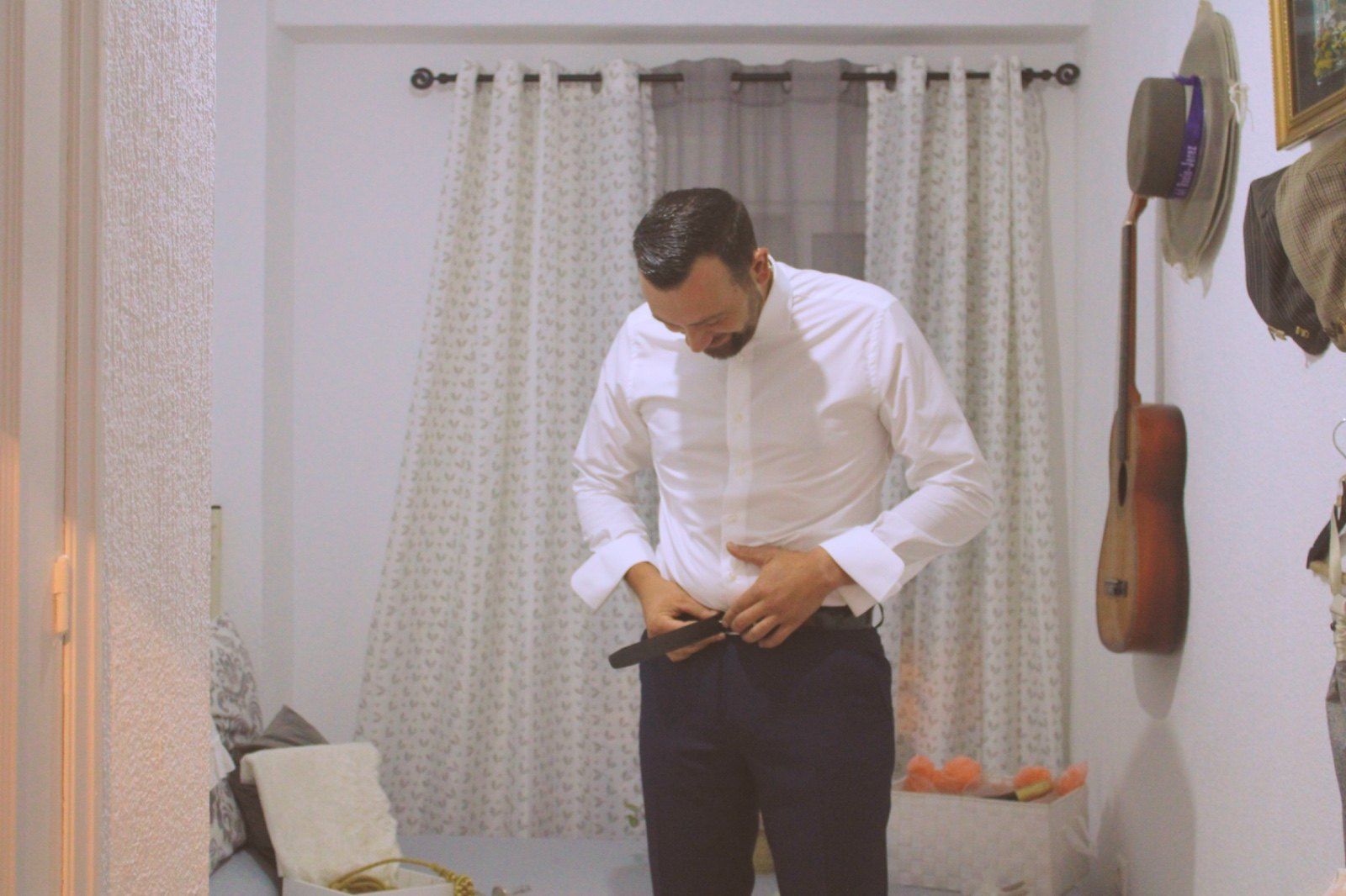 Un hombre con camisa blanca y pantalón oscuro se abrocha el cinturón en una habitación bien iluminada. 