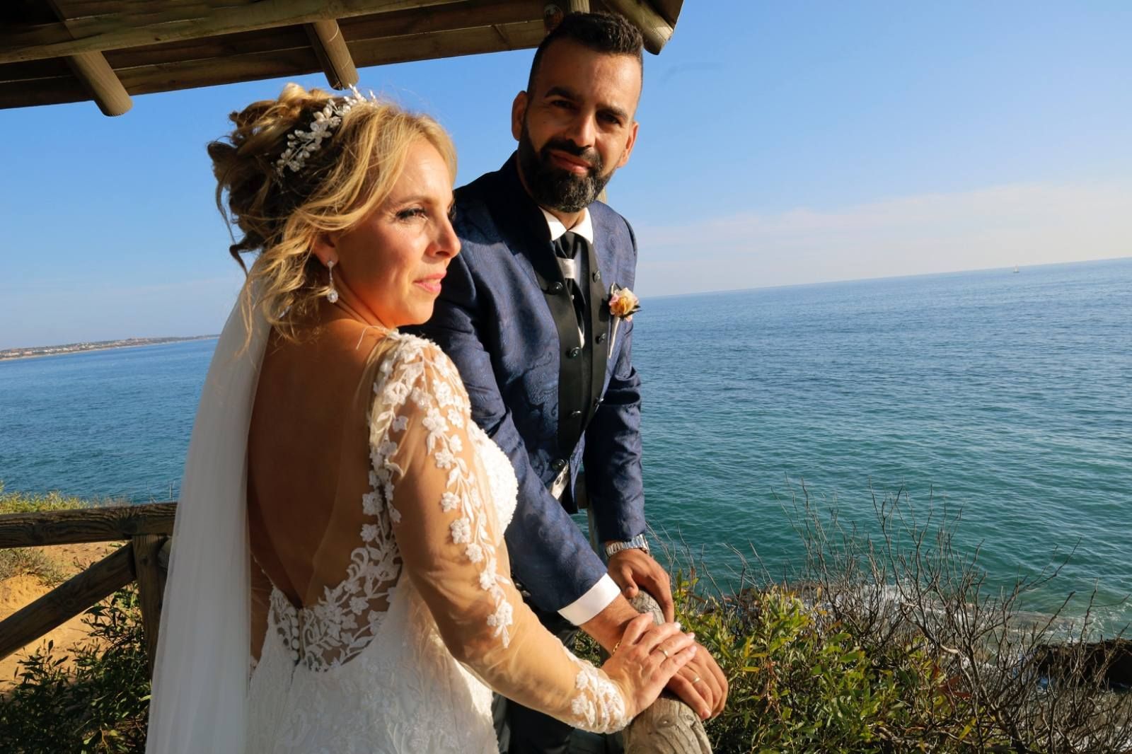 Novios en una terraza de madera con vistas al océano. La novia lleva un vestido de encaje blanco y el novio un traje azul.