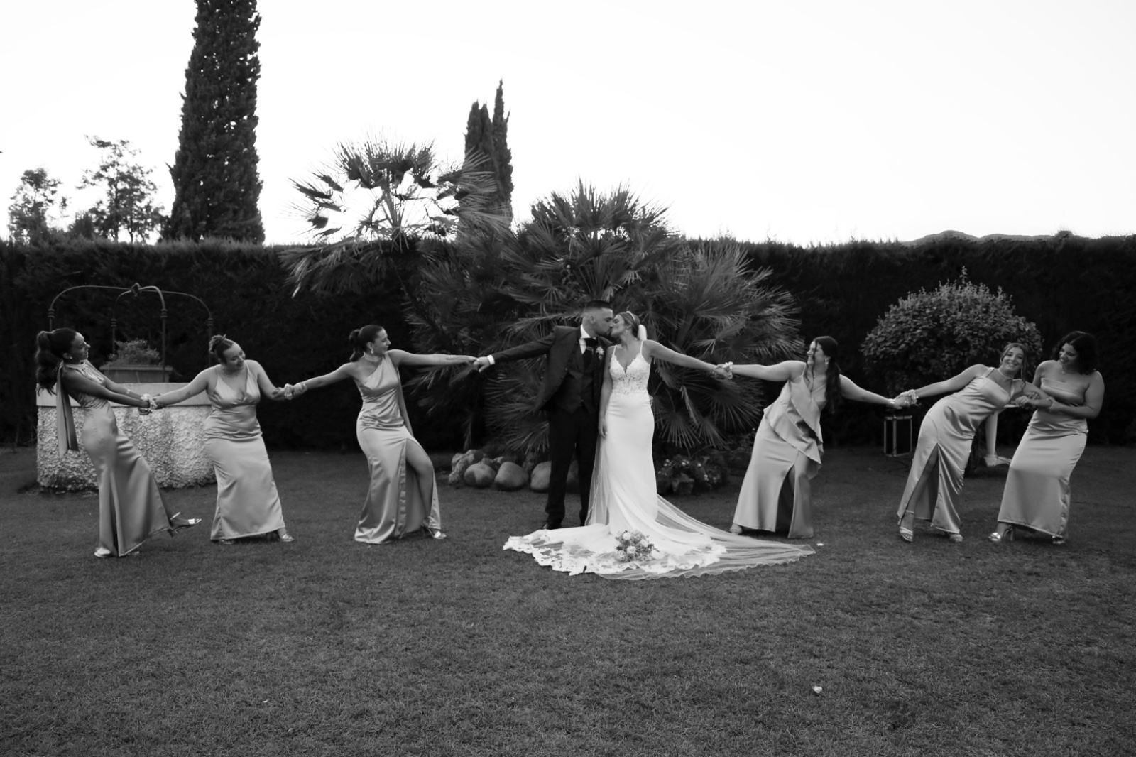 Los novios se besan rodeados de damas de honor cogidas de la mano, formando un círculo sobre el césped. Blanco y negro.