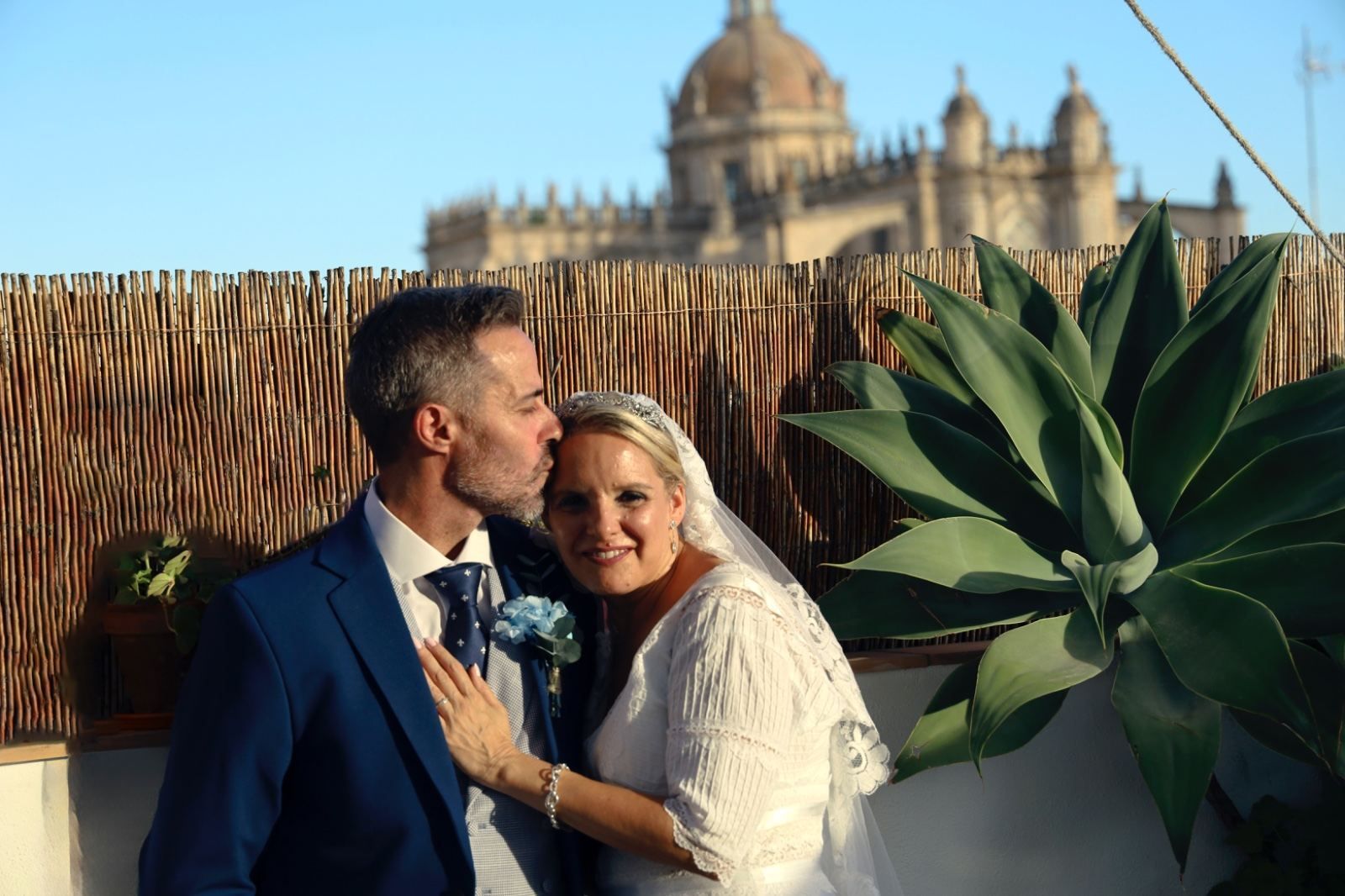 Los recién casados se abrazan en una terraza con una catedral al fondo. El hombre besa la cabeza de la mujer; ambos sonríen.
