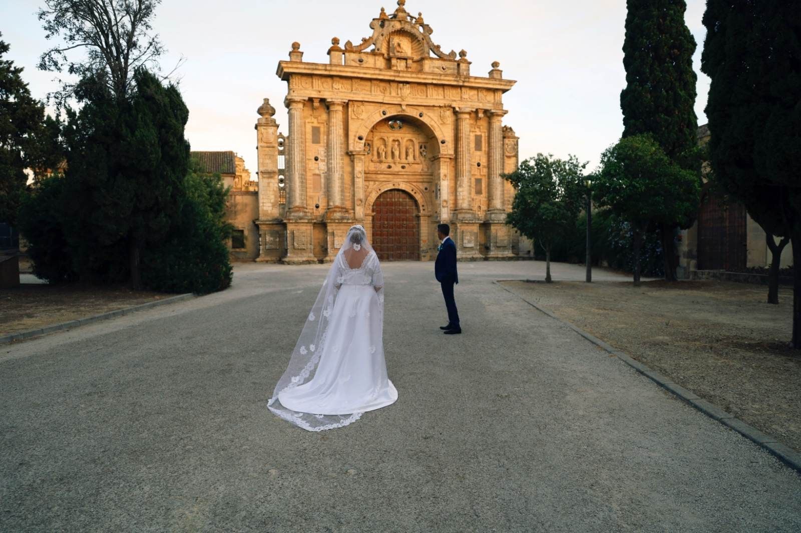 La novia con un vestido largo blanco y velo camina hacia el novio con traje frente a un gran edificio ornamentado.