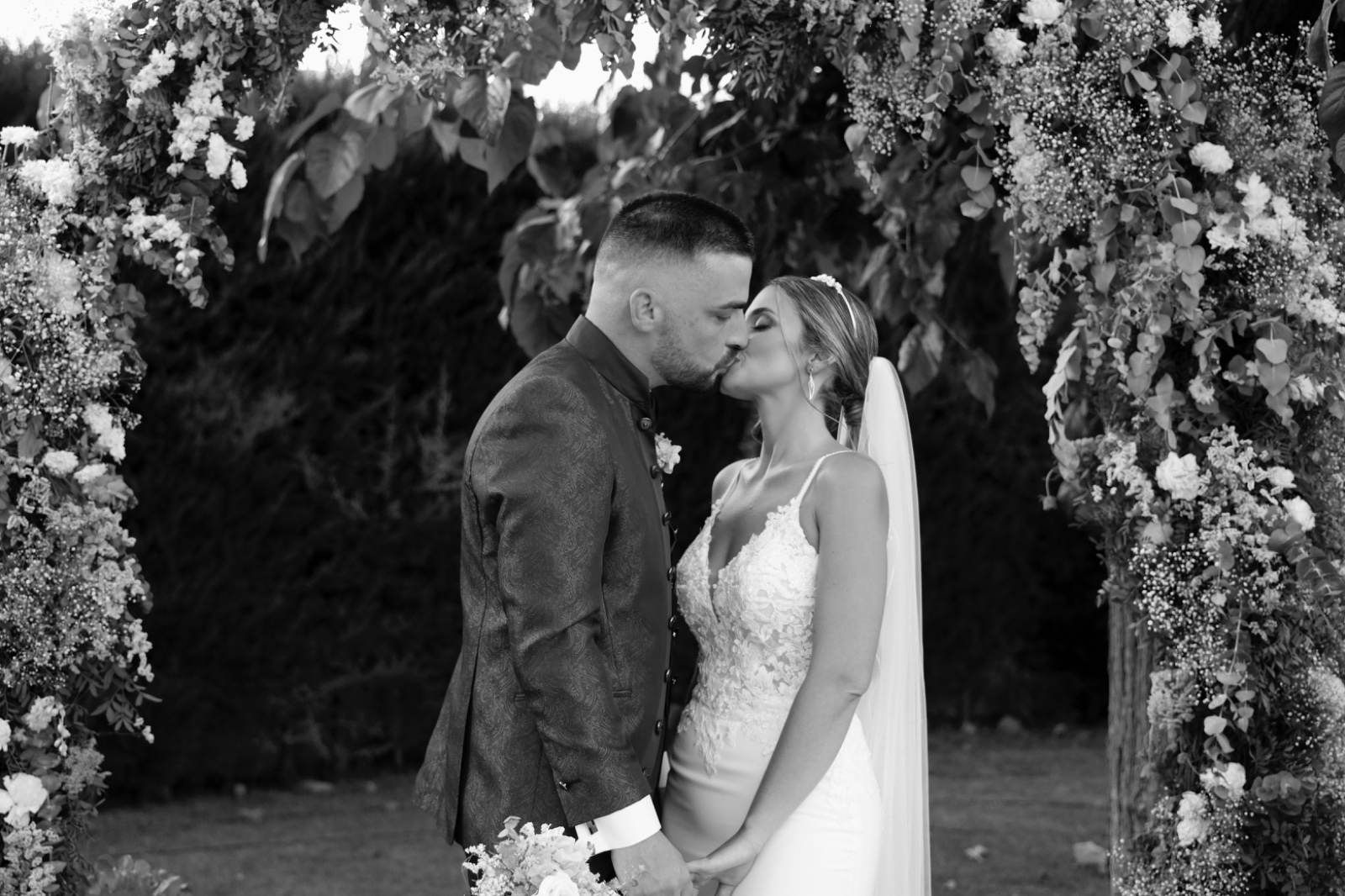 Fotografía en blanco y negro de una pareja besándose bajo un arco de flores en una boda. 