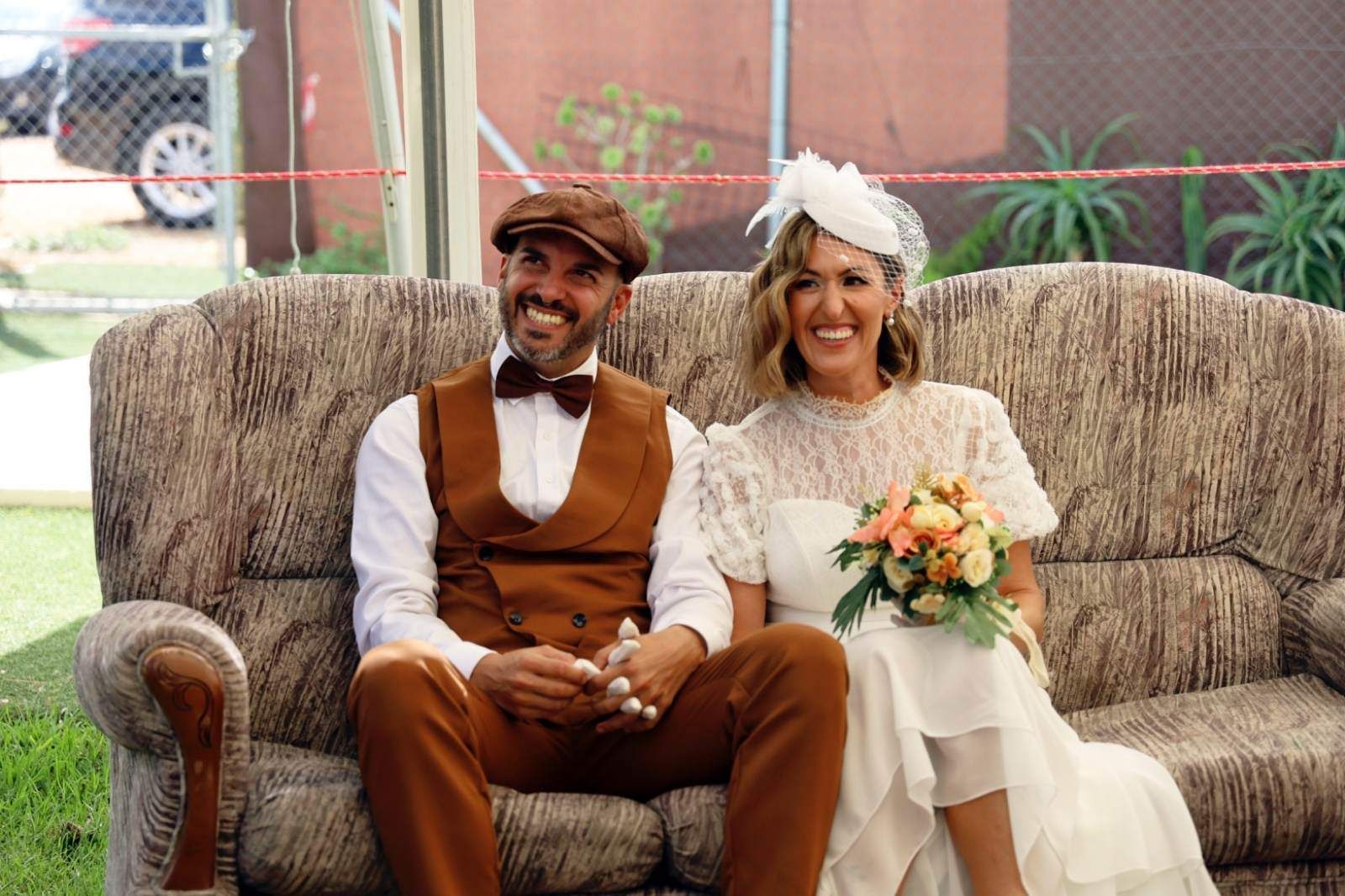 Una pareja sonriente, vestida con ropa vintage, está sentada en un sofá flora.