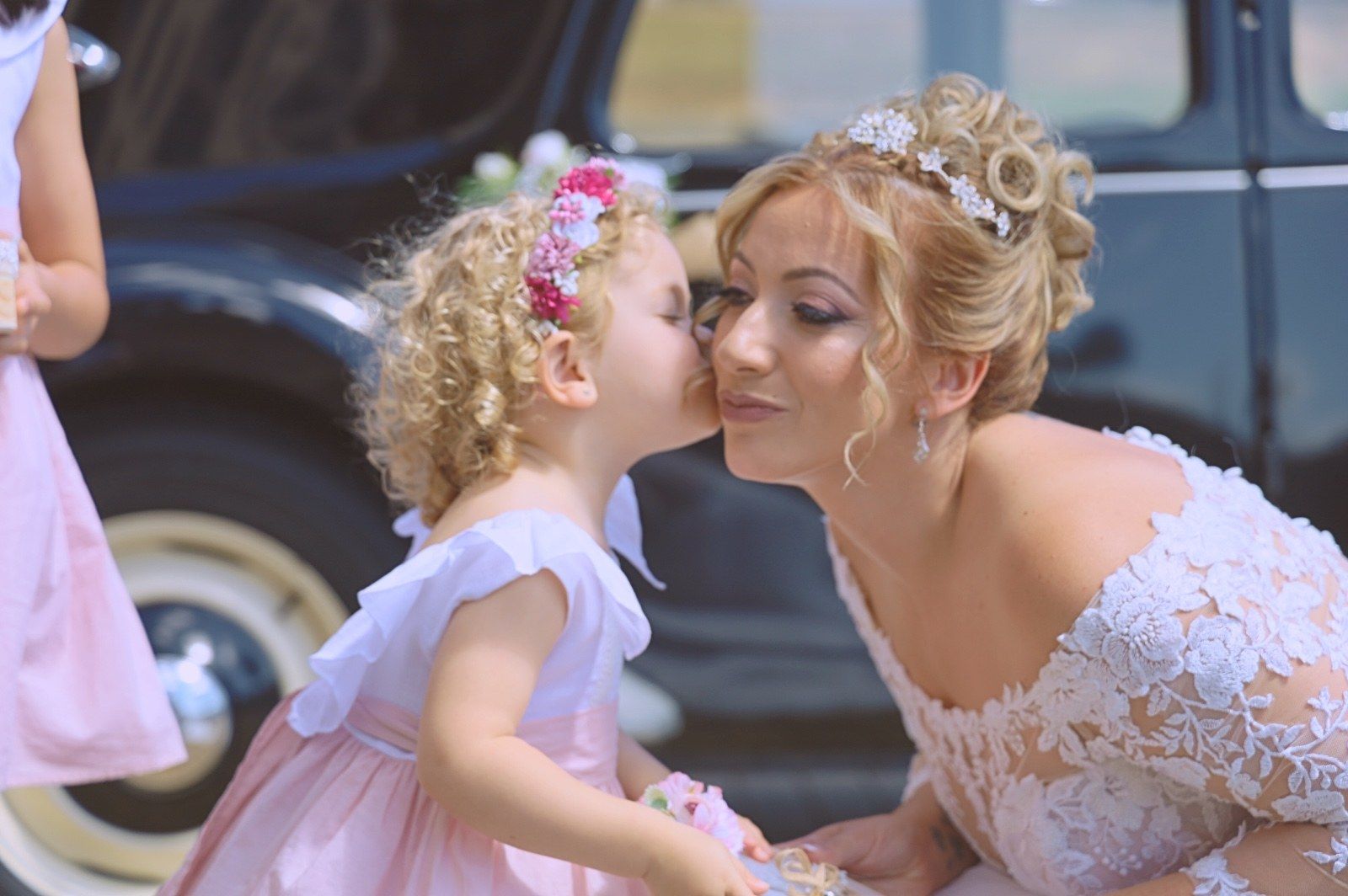 Una novia con un vestido de encaje blanco recibe un beso en la mejilla de una niña de las flores con un vestido rosa.