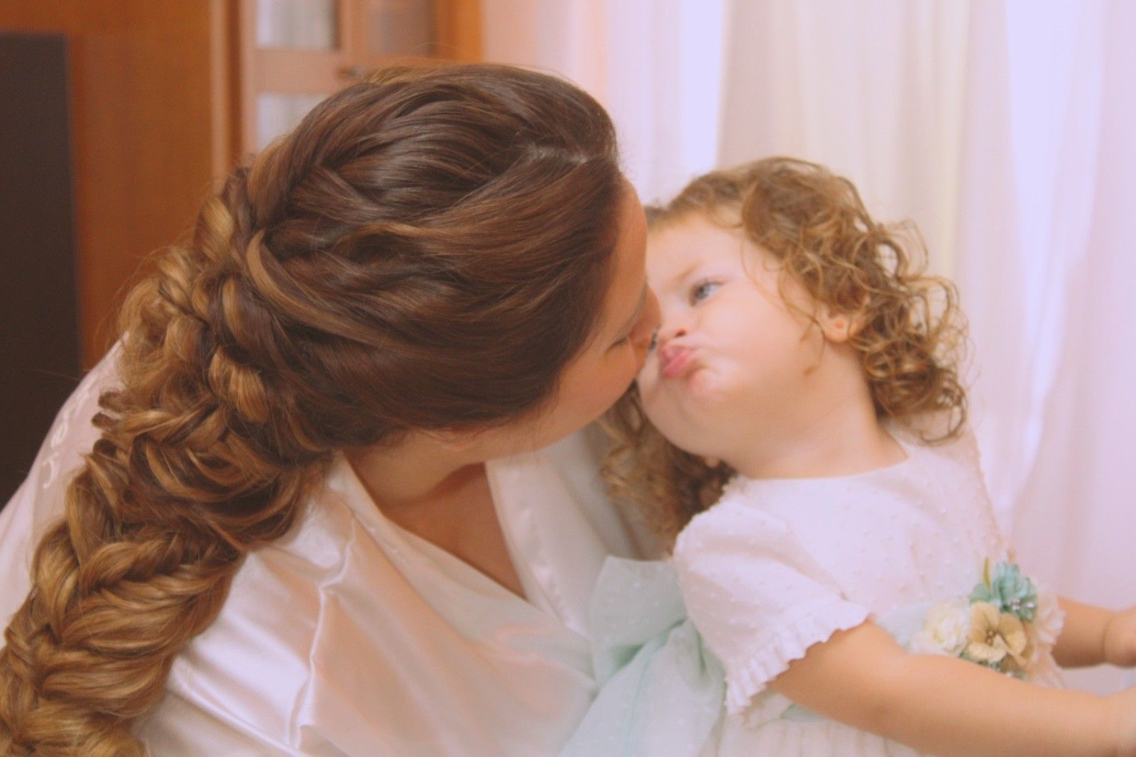 Una mujer con trenzas besa a un niño pequeño con el pelo rizado. Ambos visten de blanco, y el niño frunce el ceño.