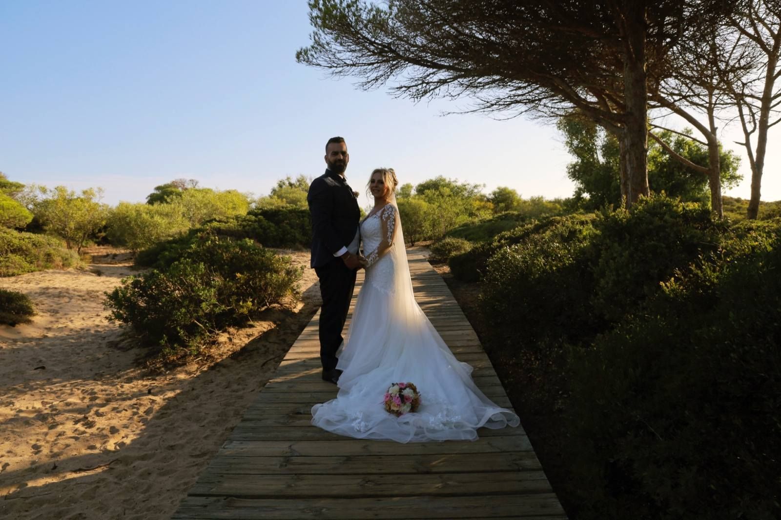 Los novios están de pie sobre una pasarela de madera en un soleado entorno al aire libre.