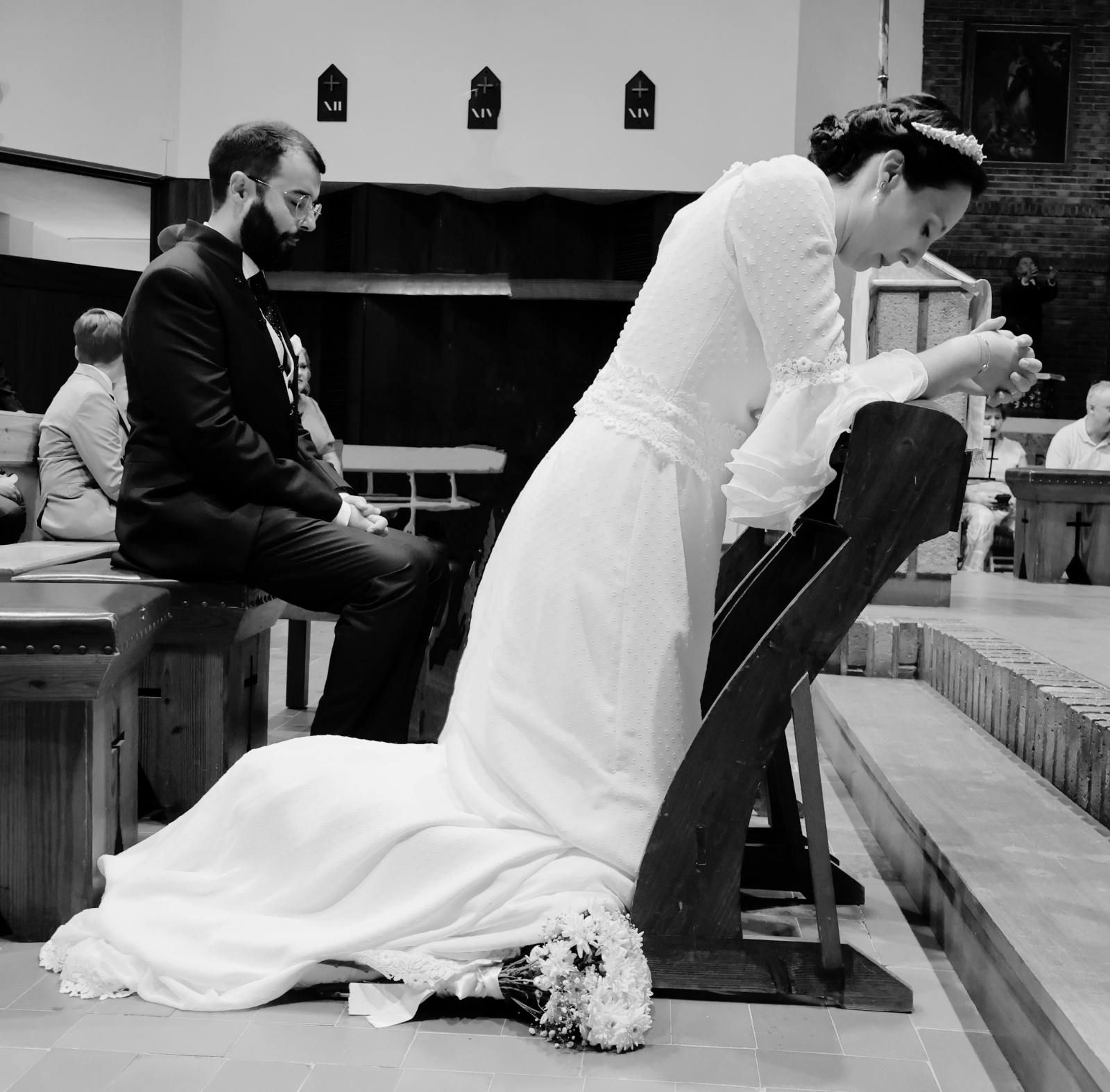 La novia con vestido blanco se arrodilla en oración en el altar de una iglesia, el novio se sienta a su lado.