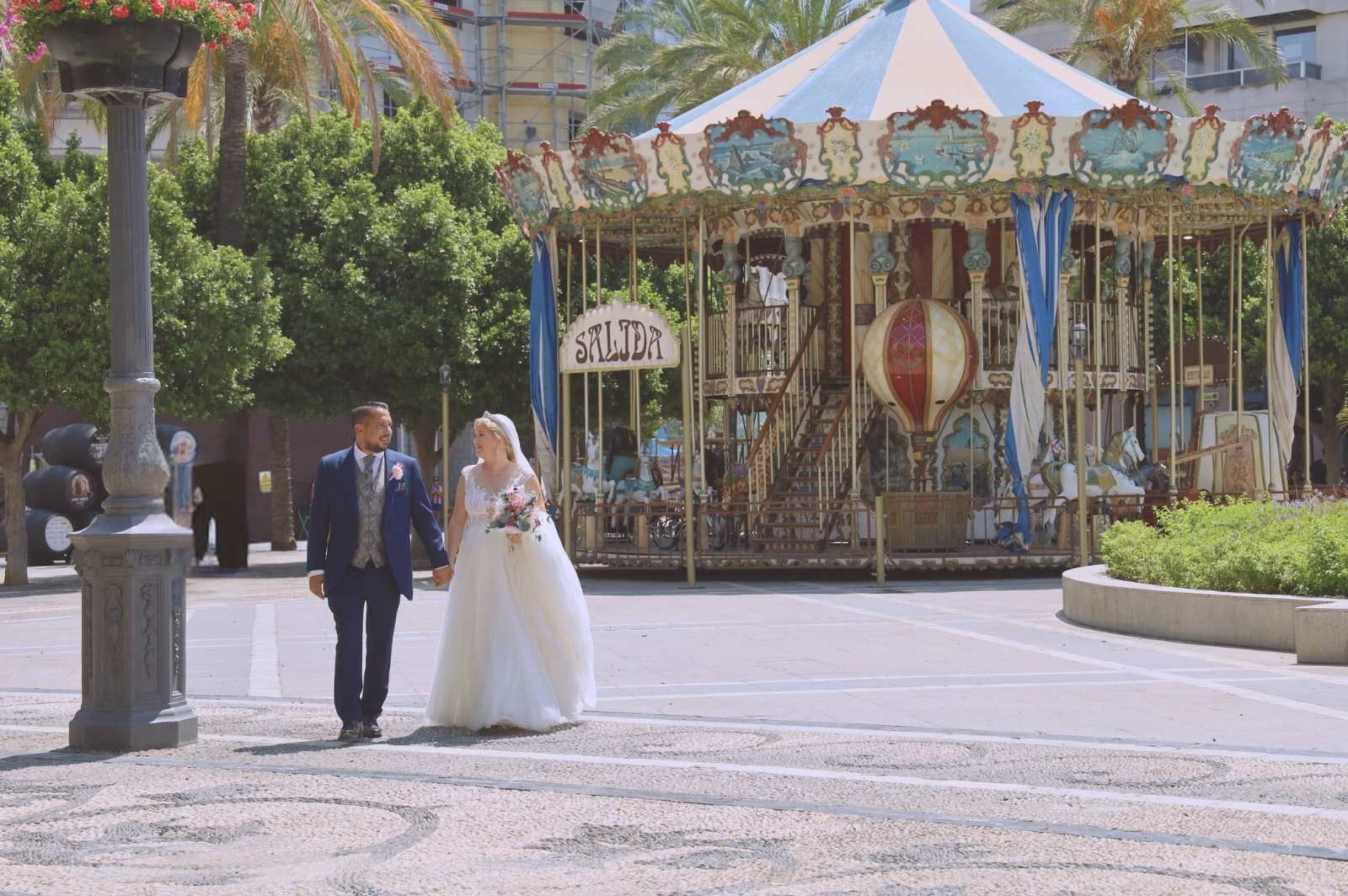 Una pareja de recién casados camina de la mano por una plaza adoquinada, con un carrusel al fondo bajo un cielo soleado.