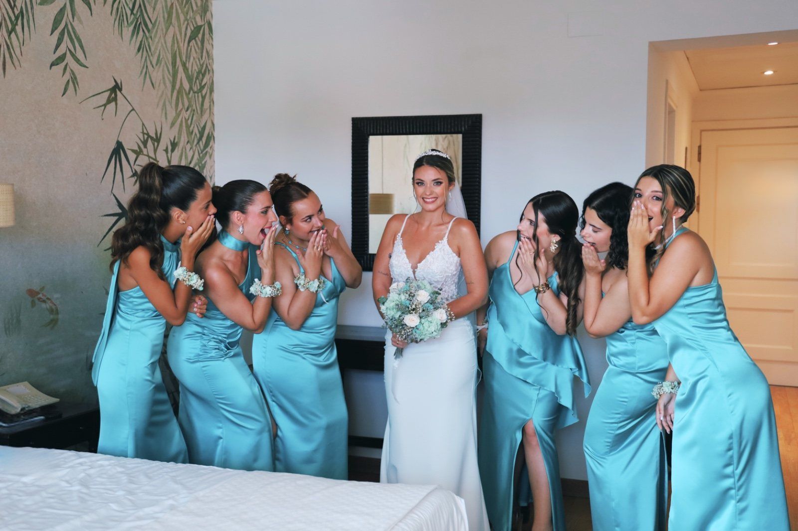 Una novia con un vestido blanco, rodeada de damas de honor con vestidos verde azulado, susurrando animadamente. 