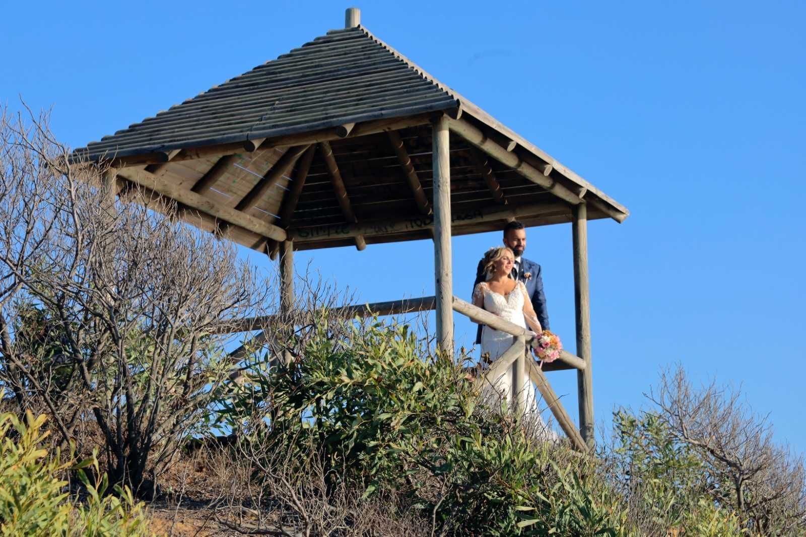 Los recién casados posan bajo un cenador de madera, con vistas a un cielo azul despejado.