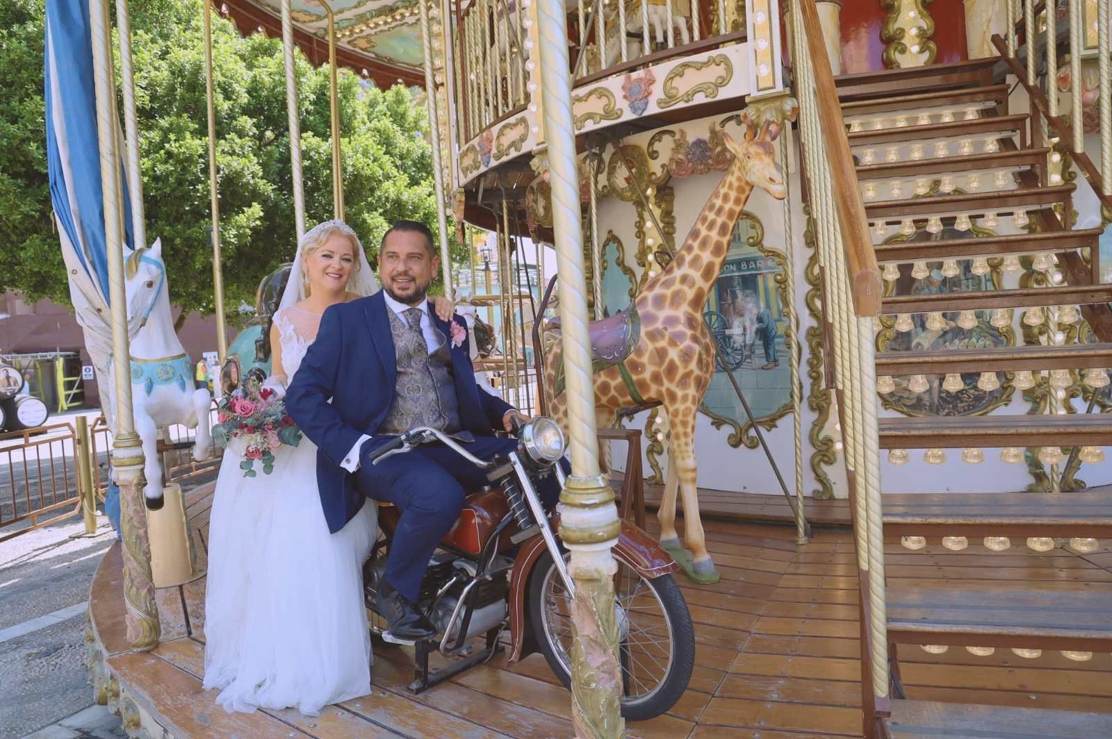 Novios en un carrusel, con el novio en una motocicleta roja. Sonríen junto a una figura de jirafa.