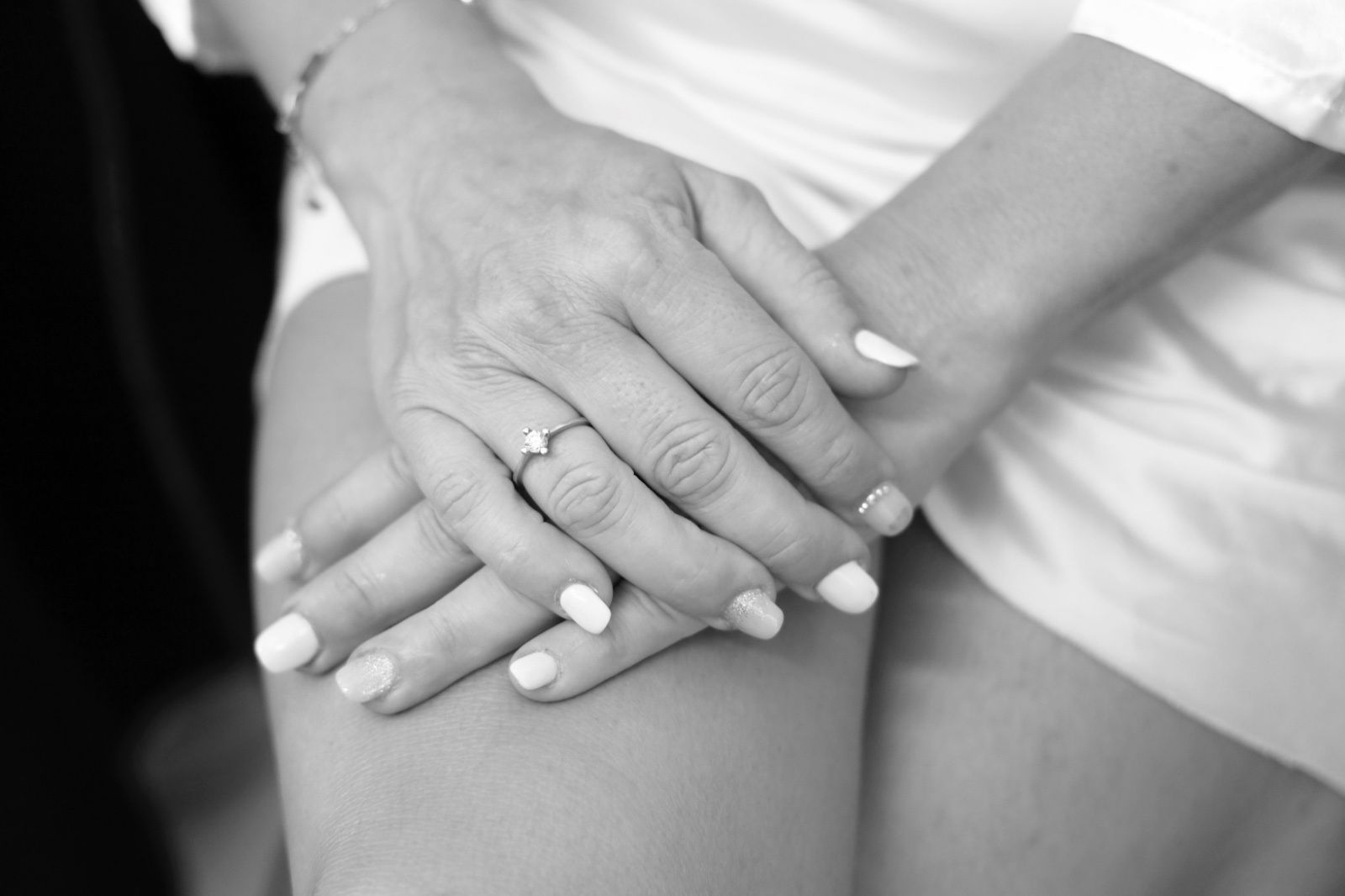 Manos de mujer cruzadas, con un anillo en un dedo y esmalte de uñas claro. Vestida con una túnica blanca, fondo desenfocado.