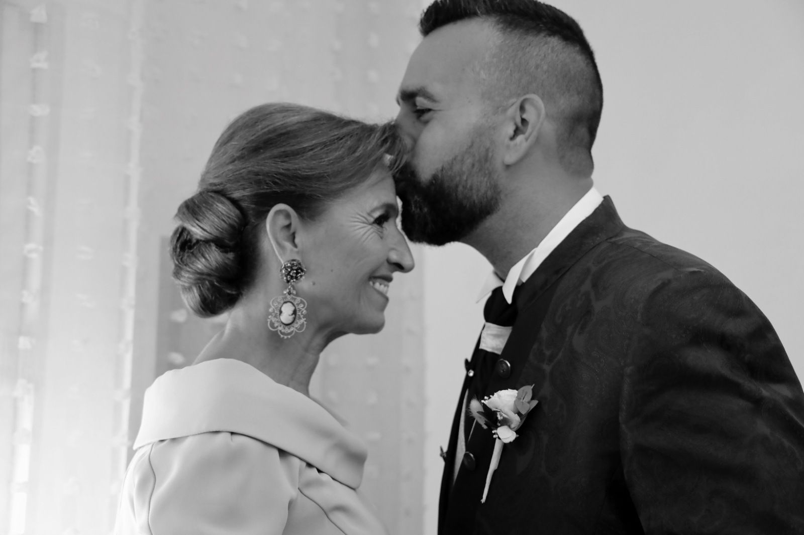 Un hombre besa la frente de una mujer en una vista cercana y de perfil. Están en una habitación iluminada. Ella sonríe.