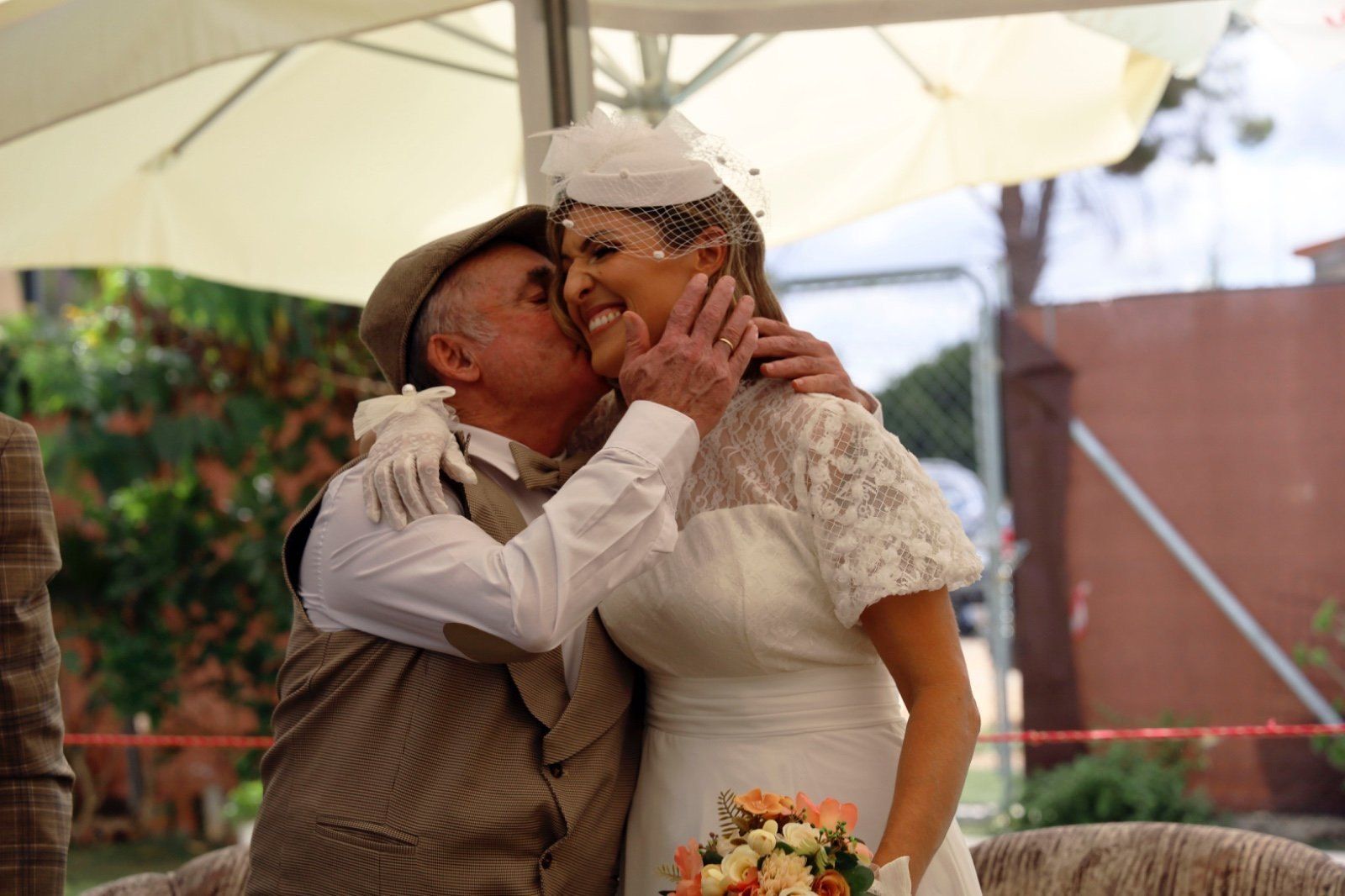Un hombre mayor besa a una novia con vestido de novia blanco y velo. Están al aire libre, bajo la luz del sol.