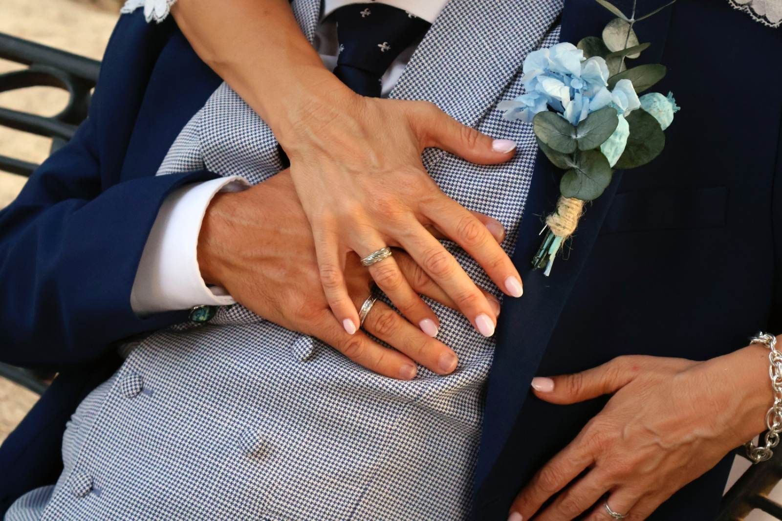 Las manos de la novia descansando sobre el pecho del novio; ambos llevan anillos de boda, traje y boutonniere.
