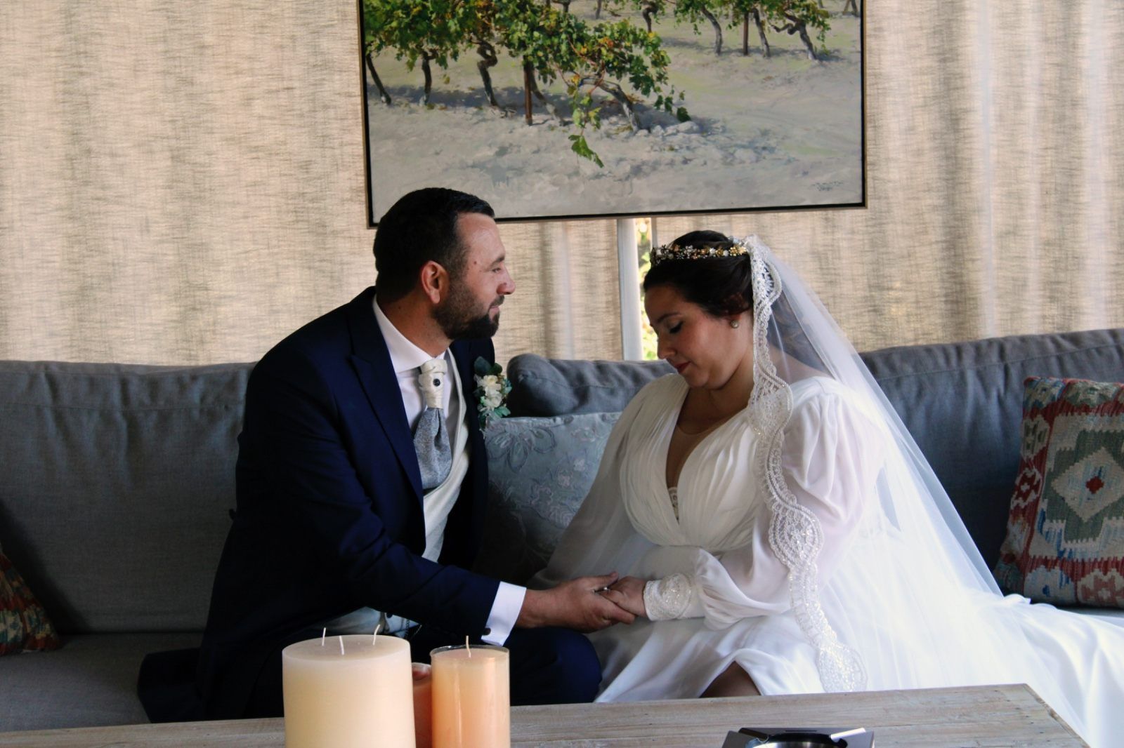 Una novia y un novio, vestidos de novia, se toman de la mano en un sofá, mirándose fijamente. 