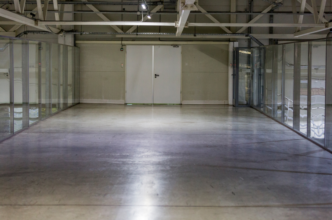 Espace industriel vide et ouvert avec sol en béton, charpente métallique et porte blanche fermée.