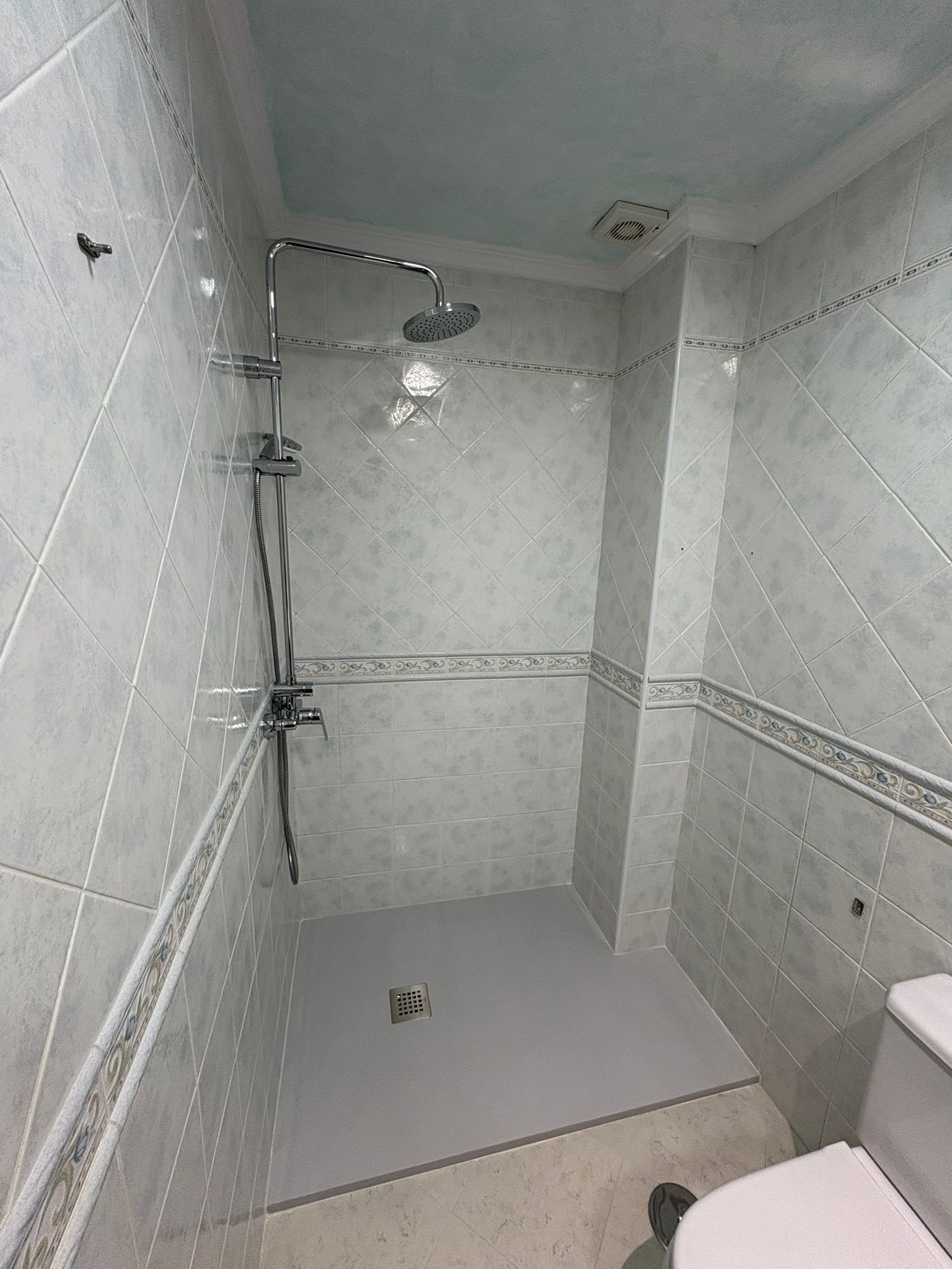 Cabina de ducha con azulejos grises y accesorios cromados, un inodoro parcialmente visible a la derecha.