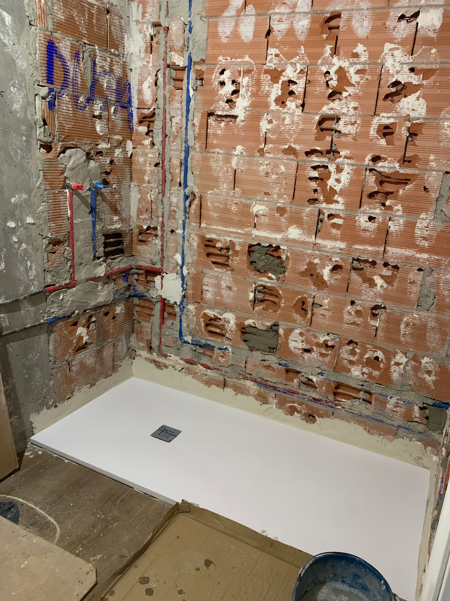 Baño en construcción con base de ducha blanca, pared de ladrillo visto y plomería.