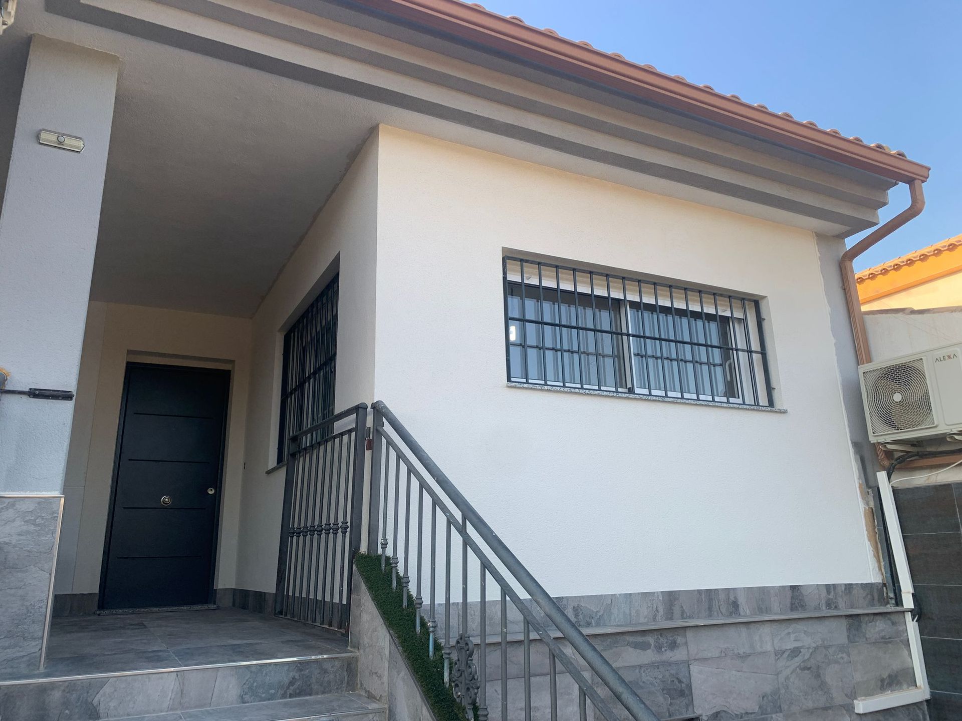 Una pequeña casa blanca con una puerta negra, ventanas enrejadas y una barandilla de metal.