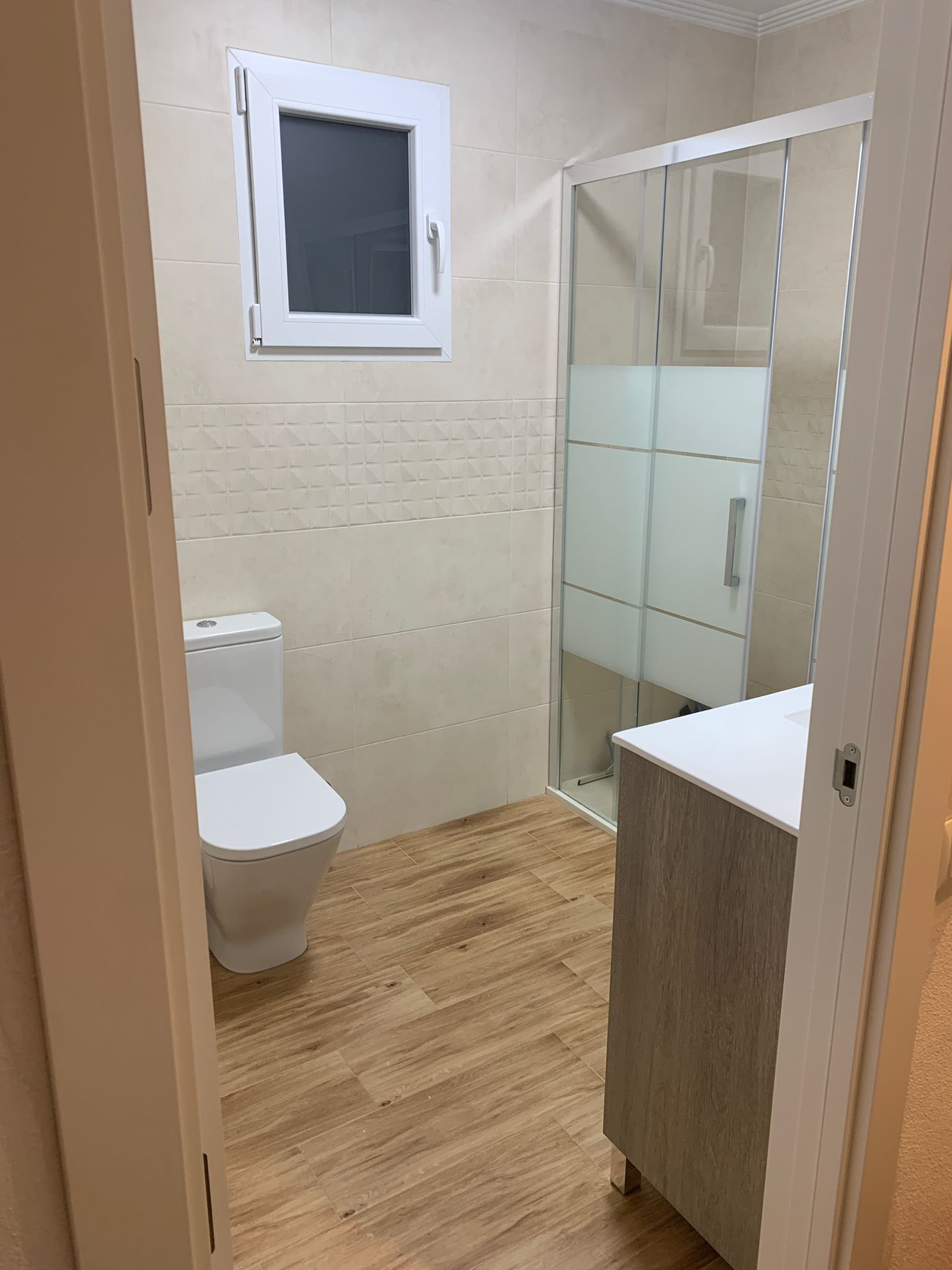 Baño con inodoro, ducha, lavabo, ventana y suelo imitación madera. Paredes beige y mampara de cristal.