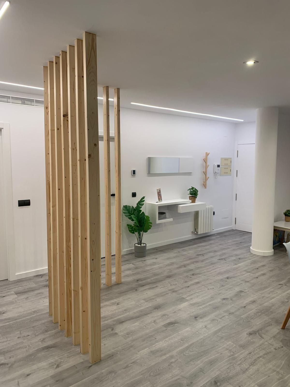 Biombo divisorio de listones de madera en un interior moderno con paredes blancas y suelo gris.