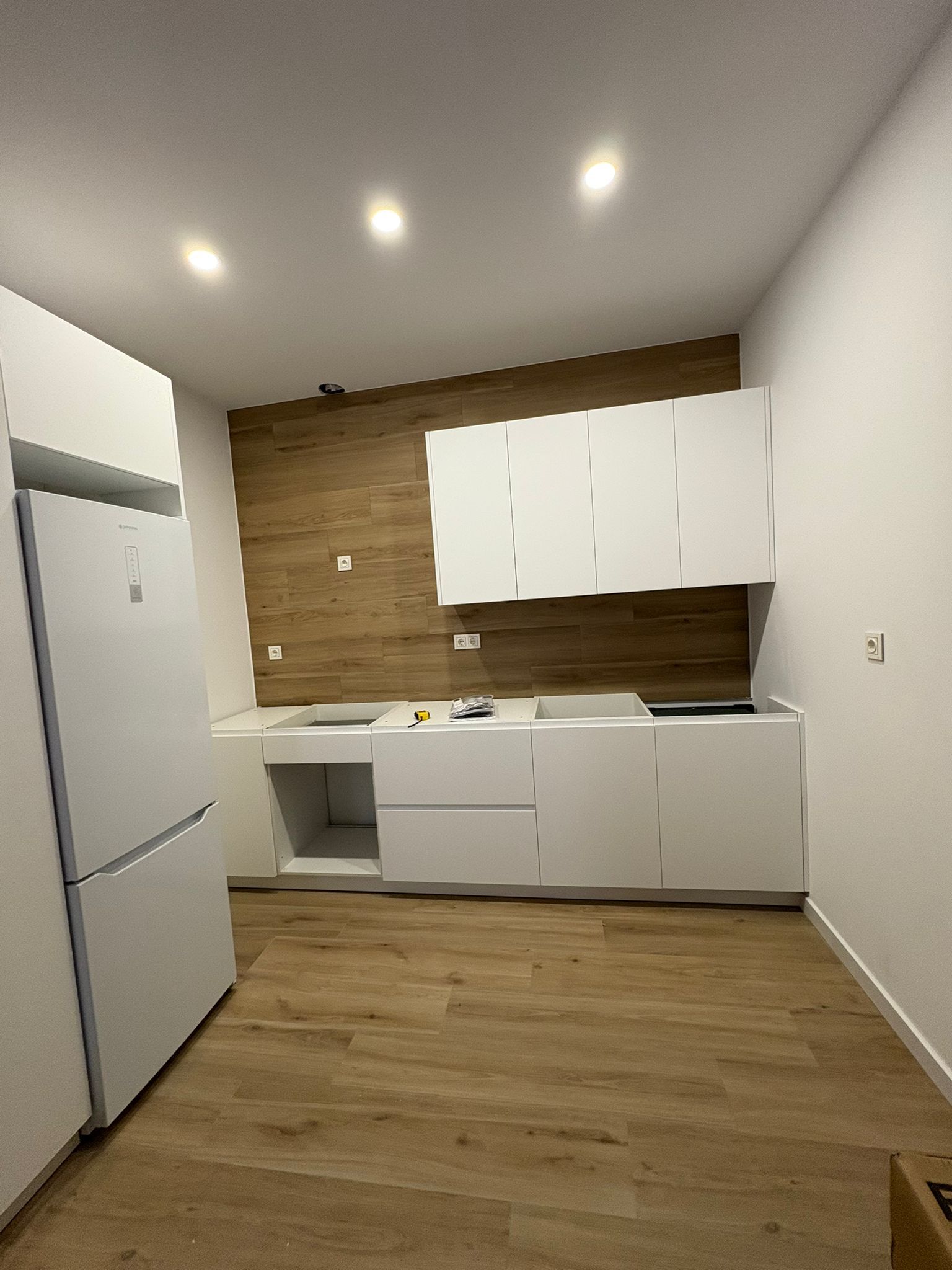 Cocina moderna con gabinetes blancos, refrigerador, pared con detalles de madera y piso de madera clara.