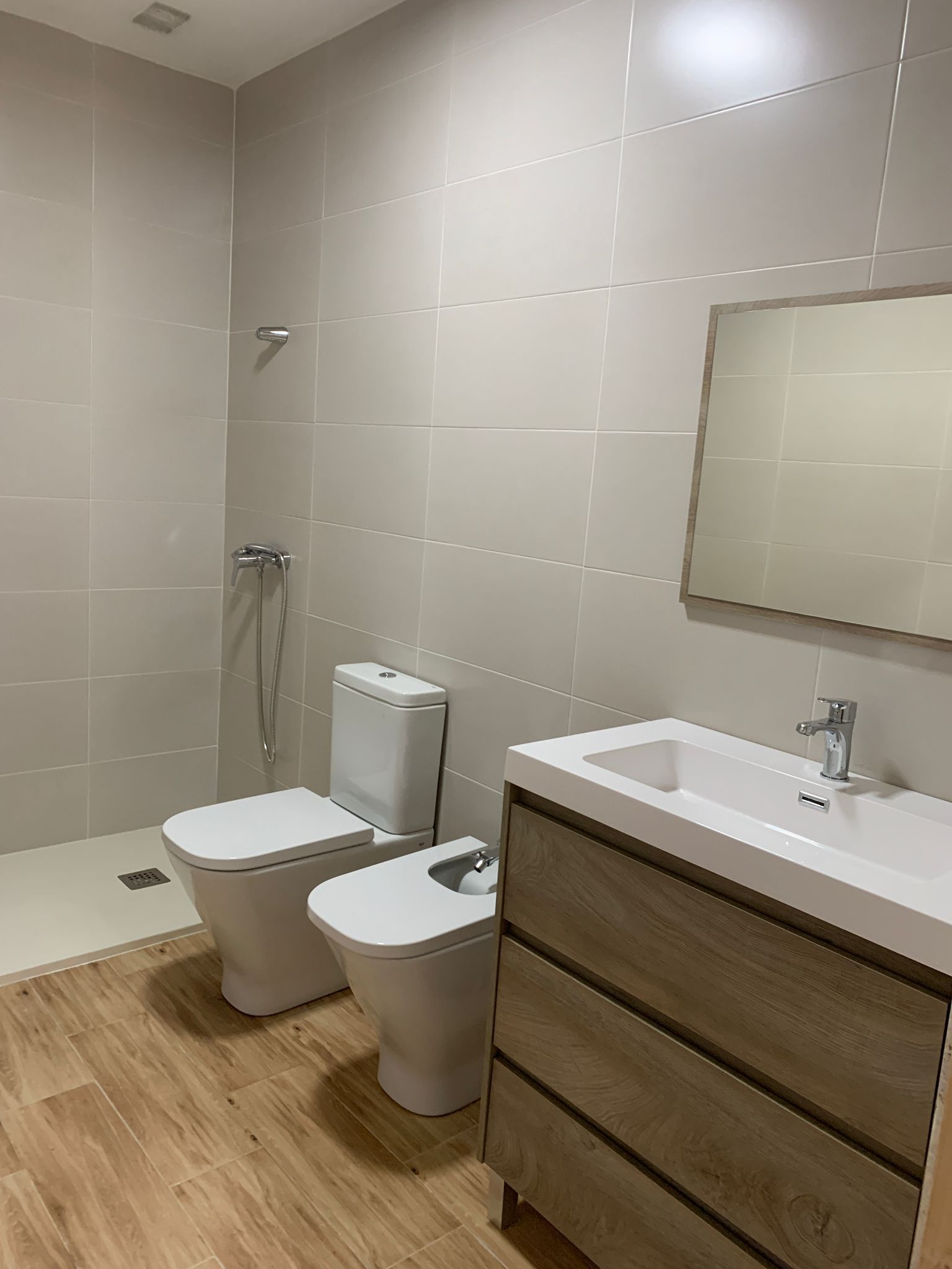 Baño con inodoro, bidé y lavabo de color blanco sobre suelo imitación madera, con paredes revestidas de azulejos de color gris claro.