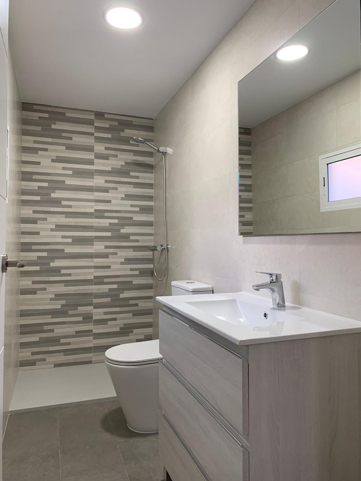 Baño moderno con azulejos grises y blancos, ducha, tocador, inodoro y espejo.