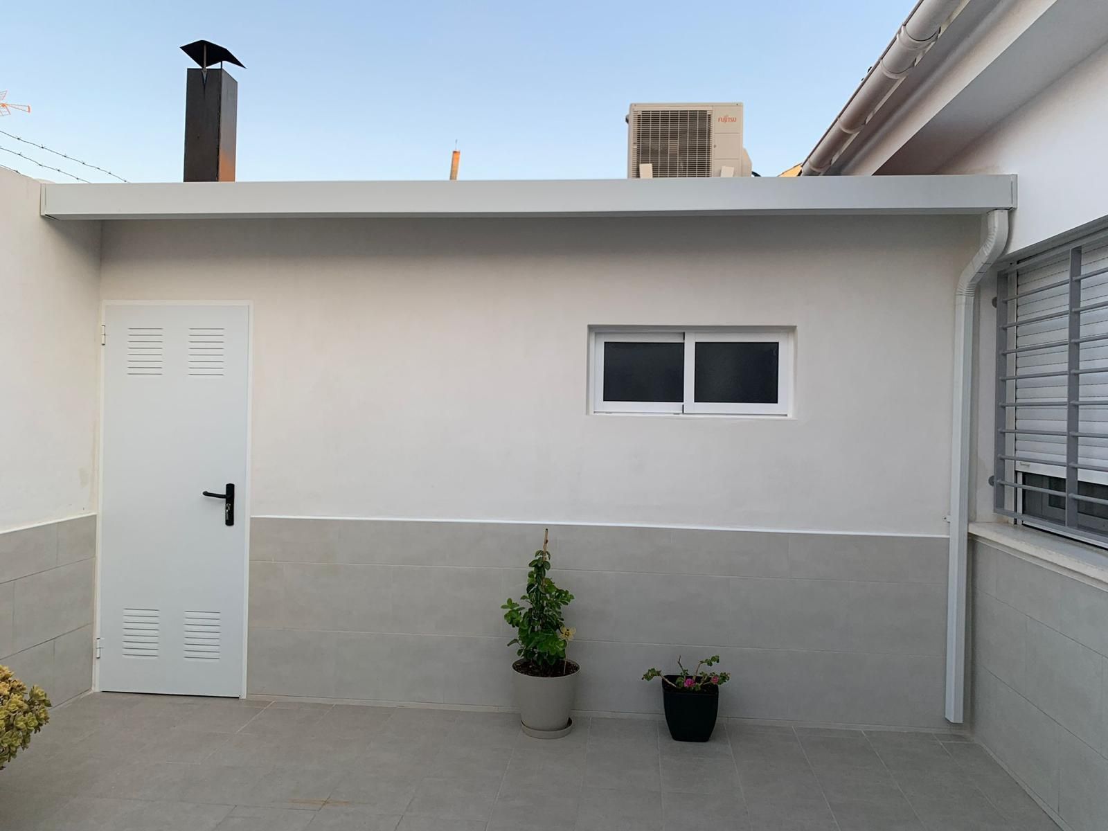 Una pared exterior blanca con una puerta, una ventana y plantas. Un pájaro posado en una chimenea.