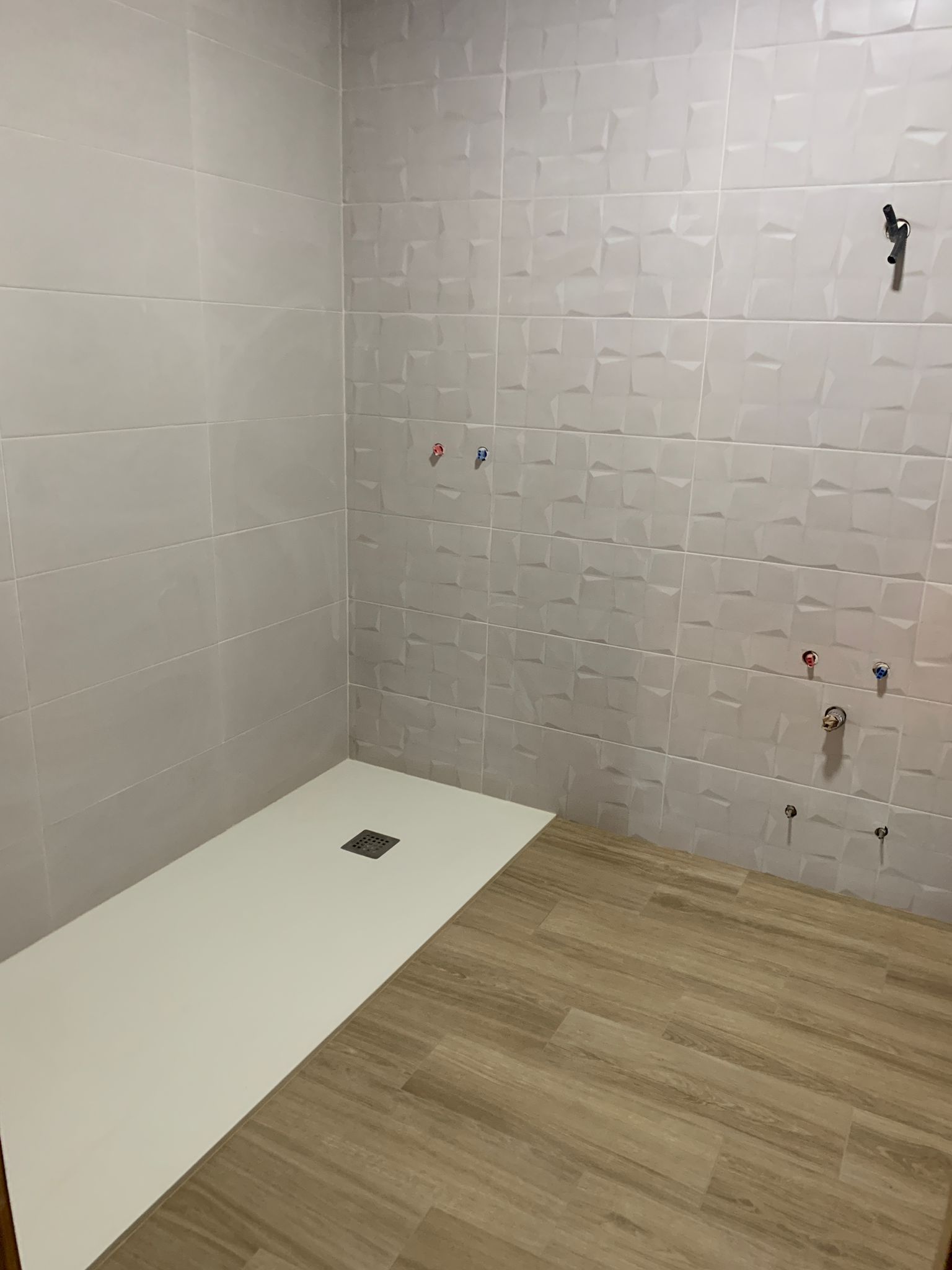 Ducha vacía con paredes de azulejos gris claro, base de ducha blanca y piso con apariencia de madera.
