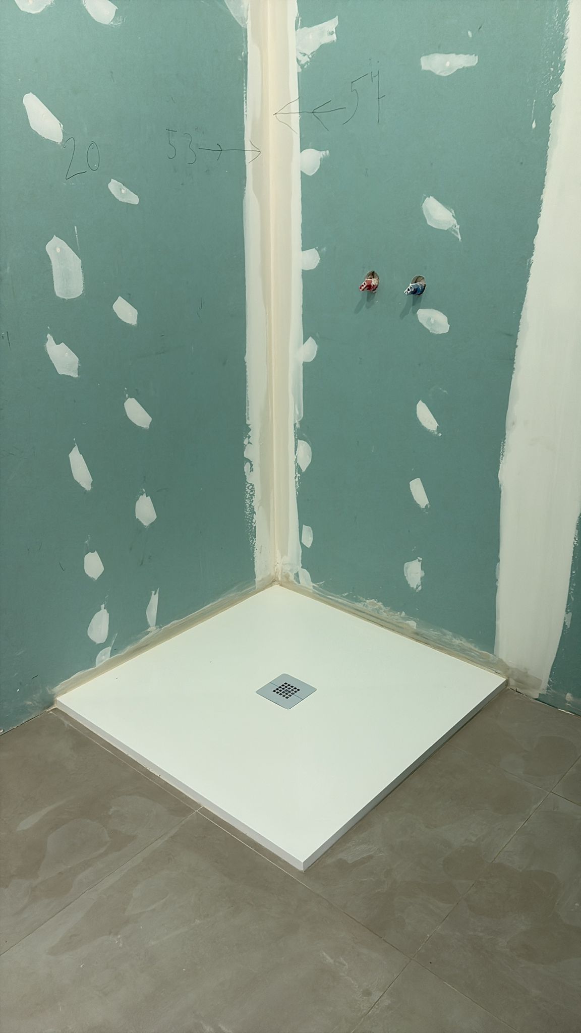 Construcción de una cabina de ducha: base de ducha blanca, paredes color aguamarina con drywall blanco, piso gris, tuberías rojas/azules.