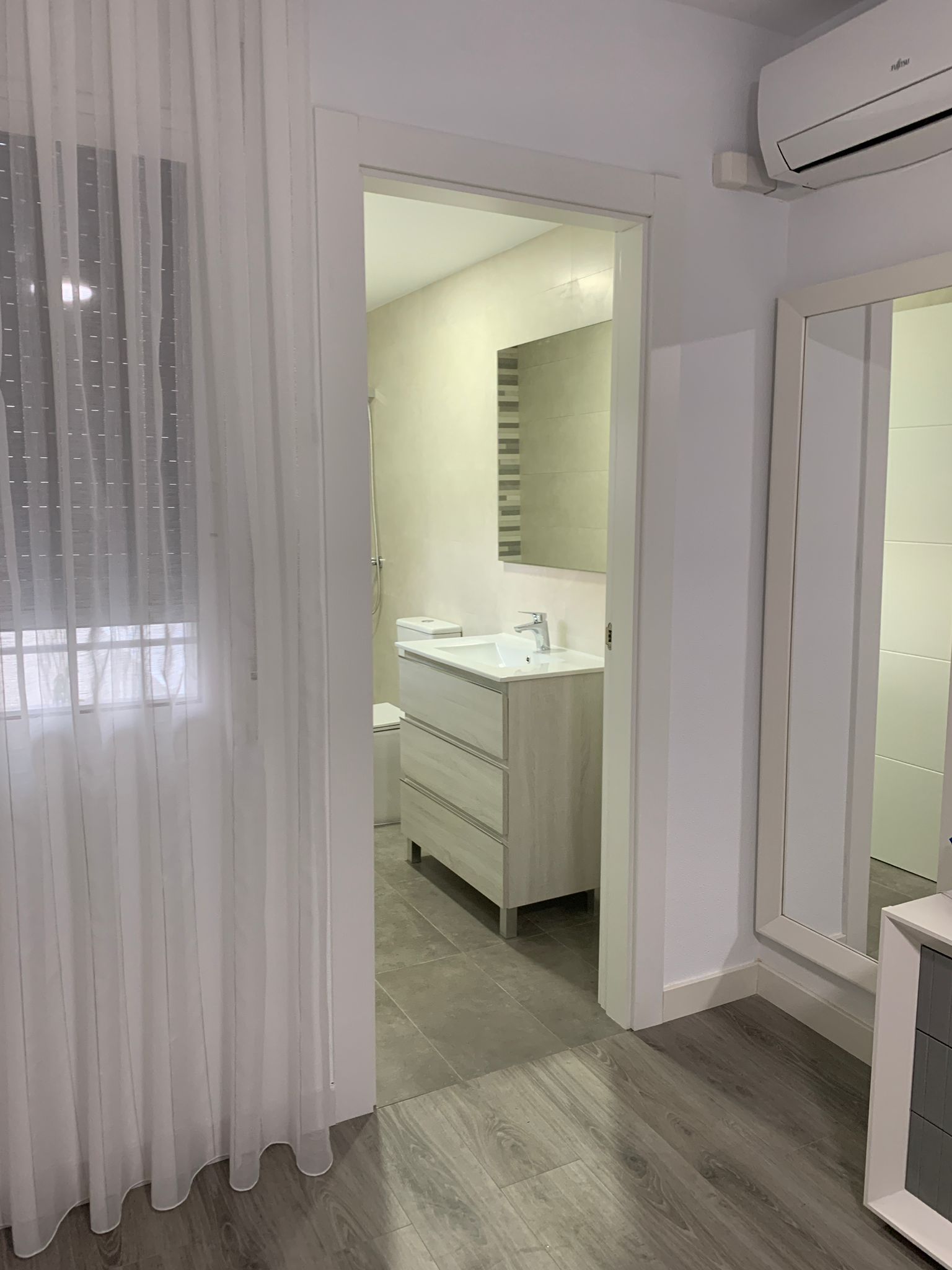 Una puerta conduce a un baño con tocador y espejo, visible desde un dormitorio con una cortina transparente.