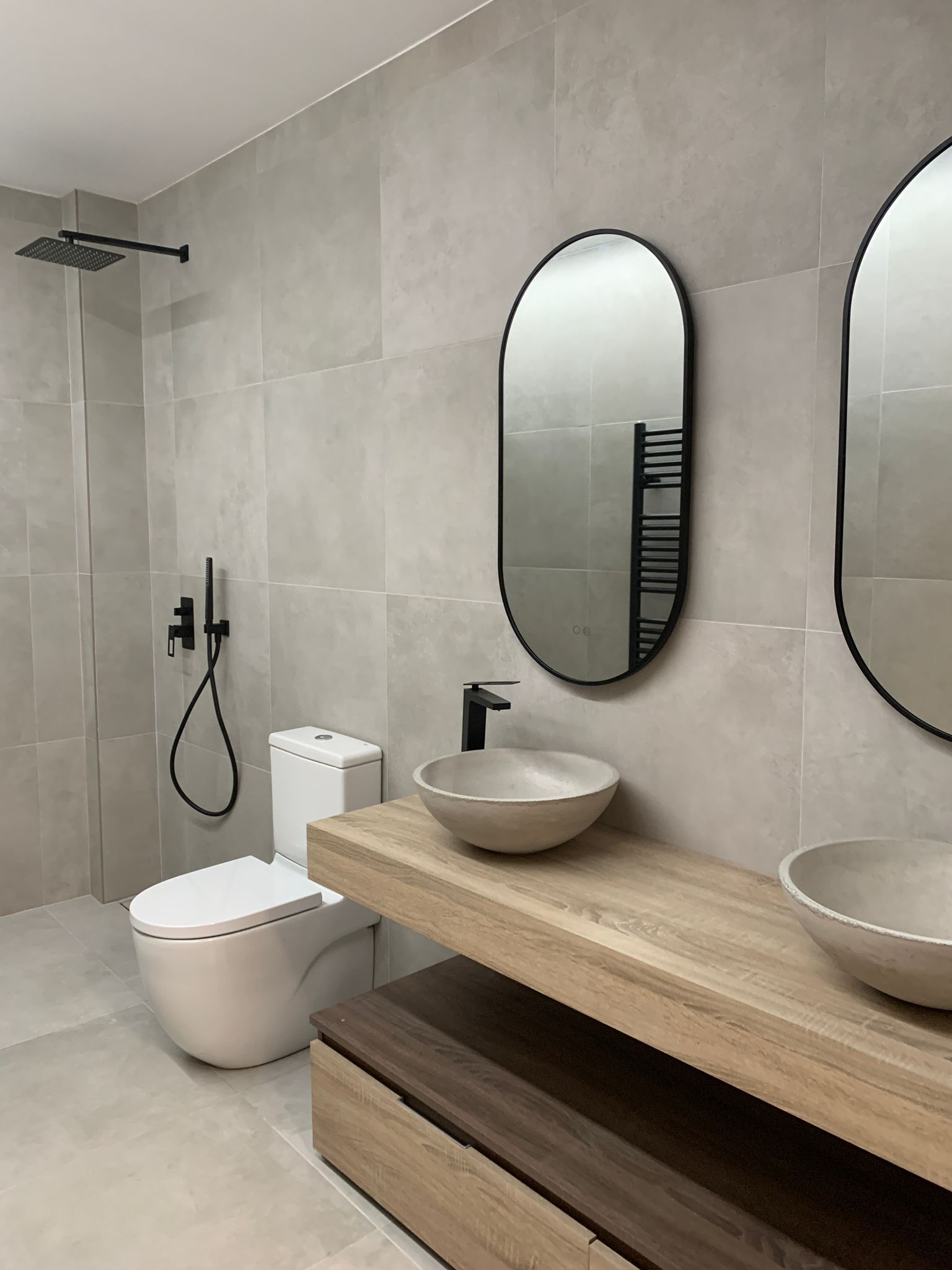 Baño moderno con paredes de azulejos grises, accesorios negros, tocador de madera clara, espejos ovalados y dos lavabos.