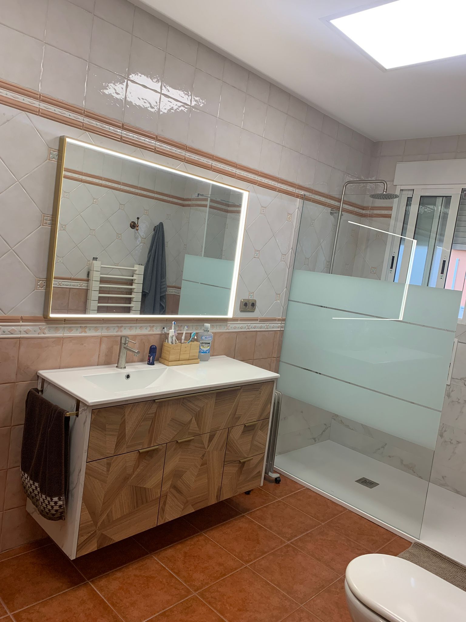 Baño con tocador, espejo grande, ducha de cristal e inodoro. Suelo de baldosas rojas y paredes en tonos neutros.