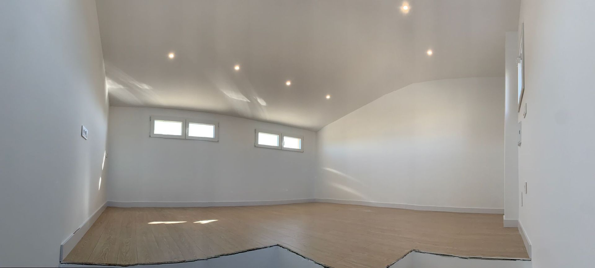 Habitación vacía, bien iluminada, con techo inclinado, piso de madera clara y ventanas pequeñas.