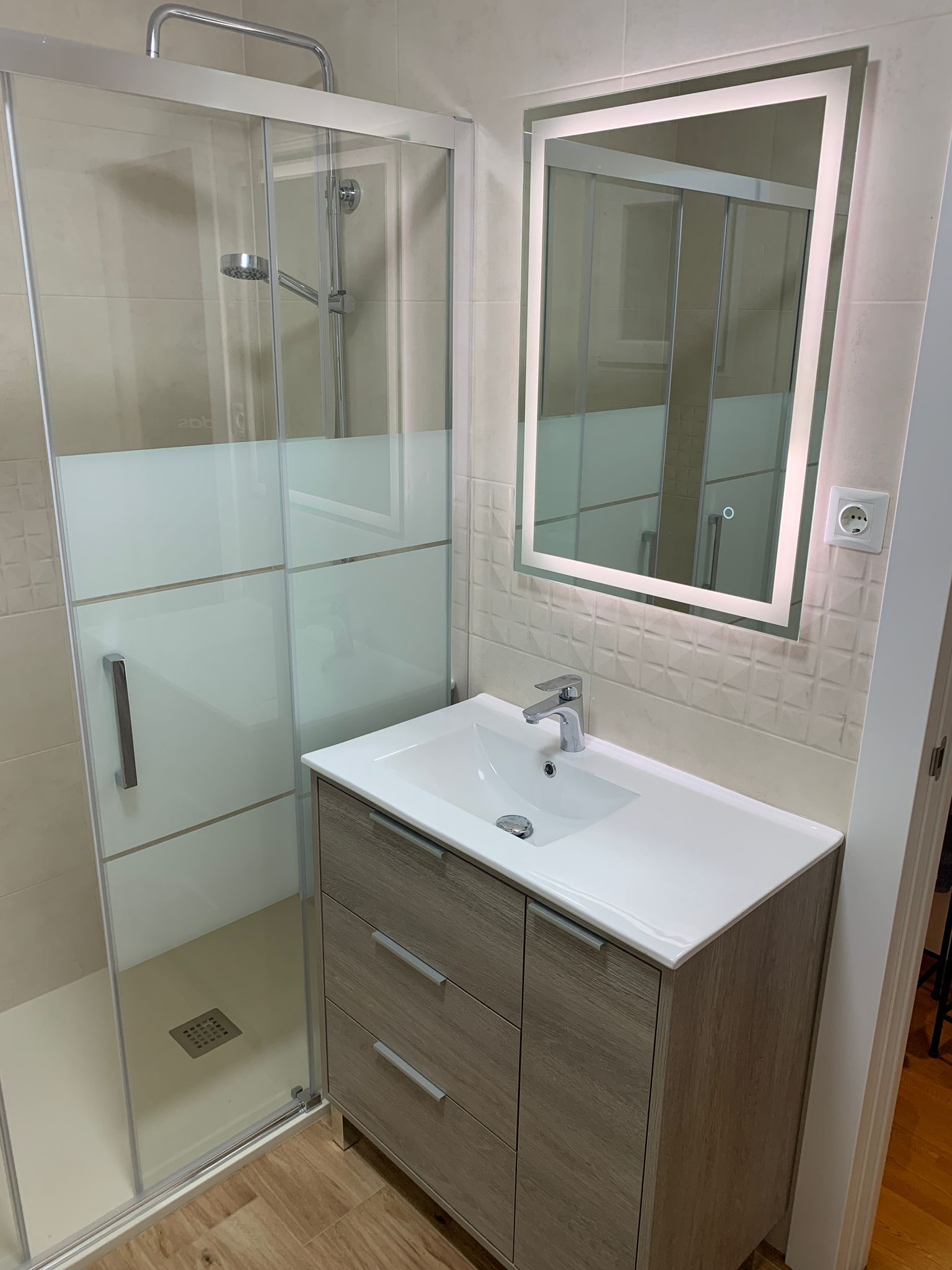 Baño con ducha, tocador y espejo iluminado. Mueble de madera clara y azulejos color canela.