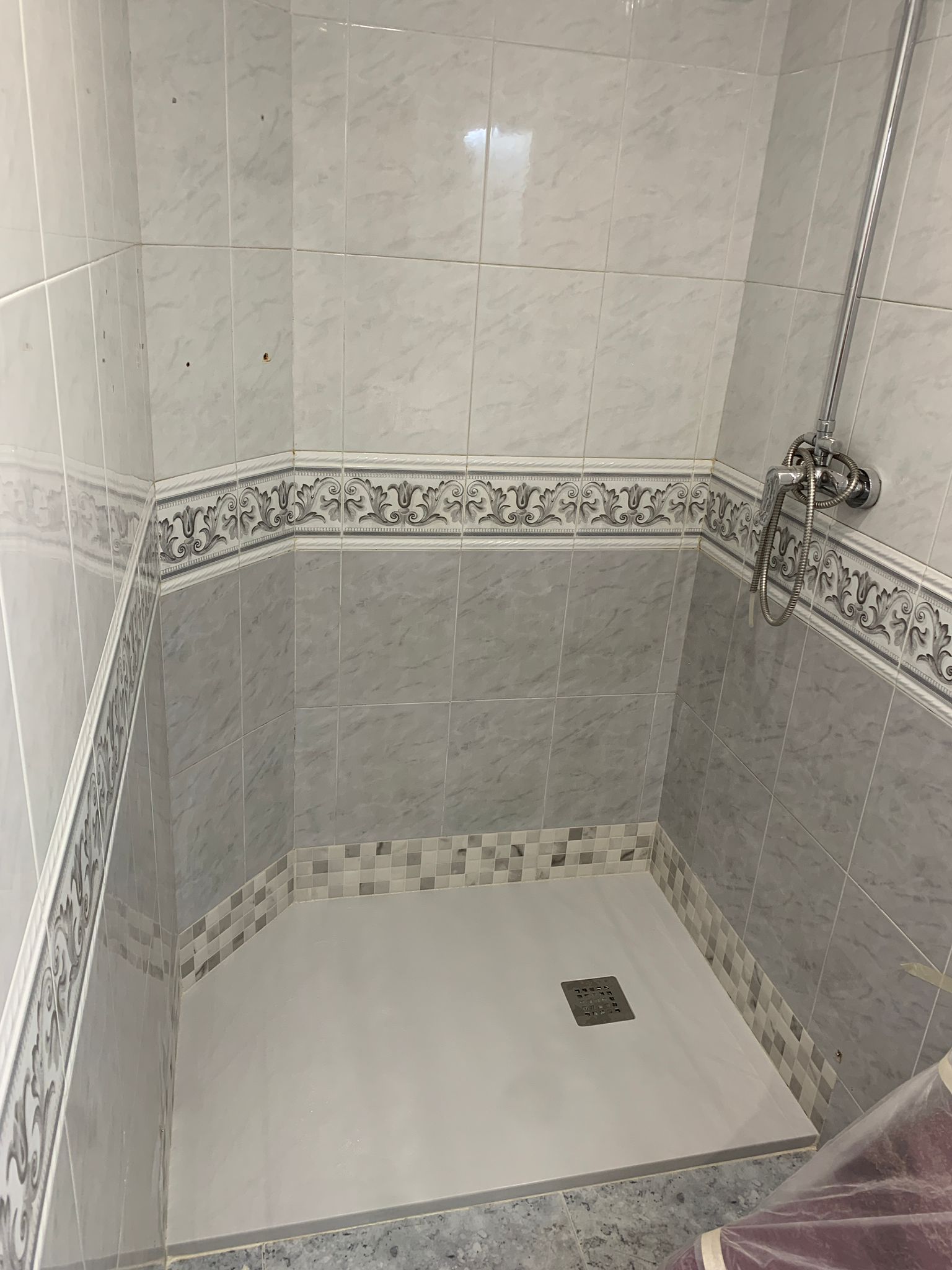 Cabina de ducha vacía con azulejos grises y blancos, borde decorativo y desagüe.
