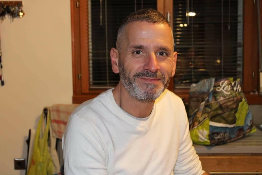 Un homme assis à l'intérieur, près d'une fenêtre, vêtu d'une chemise blanche et souriant doucement.