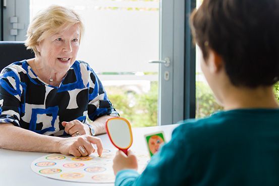 Eine Frau sitzt an einem Tisch und spricht mit einem Kind, während sie einen Spiegel hält.