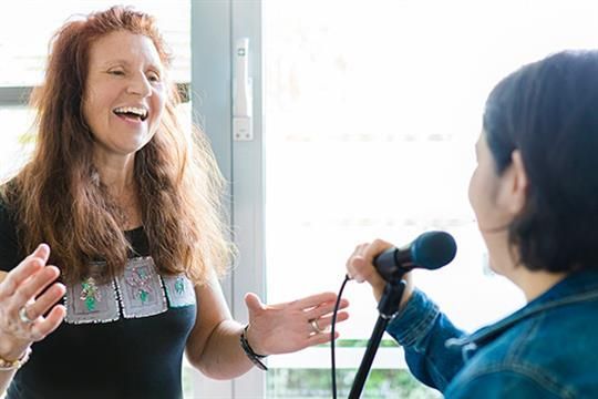 Eine Frau singt in ein Mikrofon, während eine andere Frau lacht.